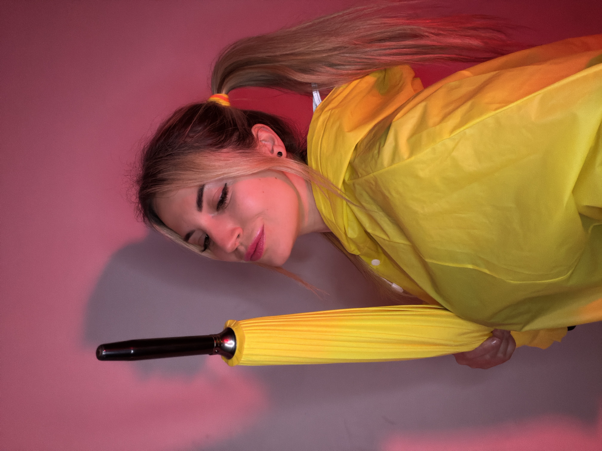 A young woman is depicted in a studio portrait, rotated 90 degrees clockwise, giving the impression she is lying down or suspended. She has long, blonde hair styled in a high ponytail, secured with a yellow and orange striped hair tie. Her eyes are gently closed or cast downwards, and a soft smile is present on her pink lips, accented by visible eyelashes. She wears a bright yellow raincoat and holds a closed yellow umbrella in her right hand, which is mostly concealed by the sleeve. A small black stud earring is visible in her ear.
The background features a smooth color gradient, shifting from a rich reddish-pink hue on the upper-left side of the frame to a lighter, more muted pink-purple on the lower-right. This lighting effect casts a warm, dramatic glow over her hair and the fabric of her raincoat. The raincoat shows some natural folds and textures. The umbrella is compact and cylindrical.
The image focuses on the subject and the interplay of the yellow and pink colors. There are no indications of the time of day, weather, or an outdoor setting. No text is visible in the frame. The general location context is Budva, Montenegro.