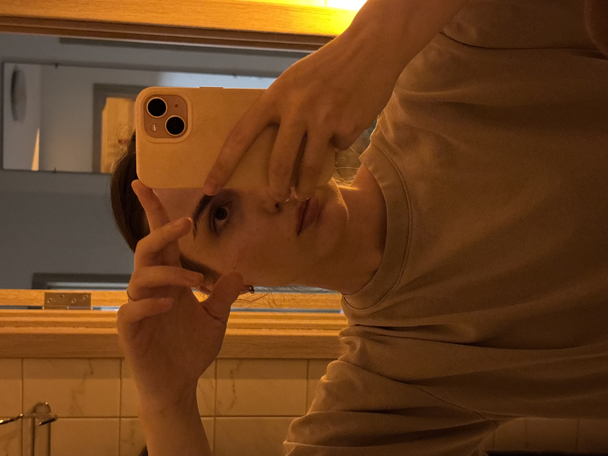 This media file is a selfie taken by a person who is partially visible. The person is holding a smartphone up to a mirror to take the photo. The phone has a light yellow case with a prominent camera bump featuring two black lenses. The person's face is visible in the reflection, showing their eyes, nose, and mouth. They appear to be looking at the camera.
The person is wearing a light brown, possibly tan or beige, t-shirt. The fabric of the shirt shows some texture and folds, suggesting it's a soft material. The background of the selfie is a bathroom setting. The mirror reflects a light-colored tiled wall, and a wooden shelf or ledge is visible above the tiles. The lighting in the scene is warm and artificial, possibly from overhead lights or a vanity lamp, casting a golden hue. There's also a hint of a darker, possibly gray wall or fixture reflected in the mirror further in the background. The overall mood seems casual and personal, typical of a mirror selfie. There is no visible text in the media file. The time of day is not explicitly indicated but the artificial lighting suggests it could be evening or night.