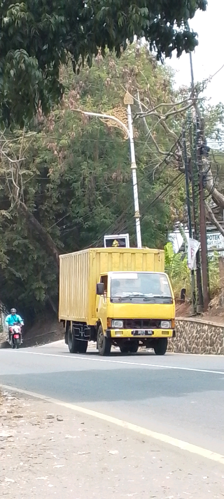 A yellow box truck is driving down a road in Cilaku, Indonesia. The truck is the primary subject, with its front facing the camera. A person on a motorcycle, wearing a blue shirt and a helmet, is visible on the left side of the road, also moving forward. The road is paved and appears to be a two-lane street. Trees and foliage line the right side of the road, with some branches extending over the street. A stone wall is visible behind the truck on the right. Electrical poles with wires are present in the background. The sky is overcast.