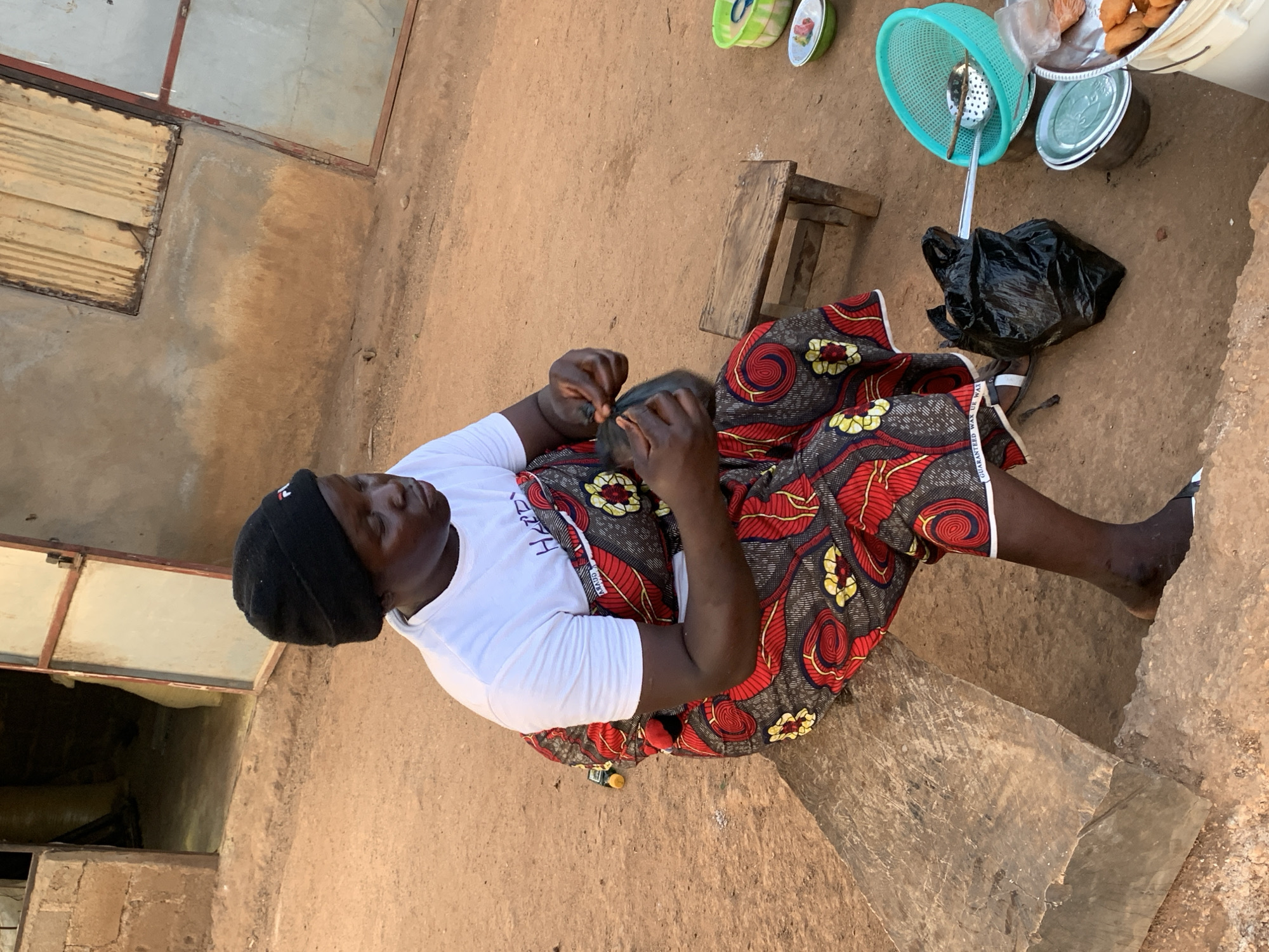 A woman wearing a black beanie, white t-shirt, and red and black patterned skirt sits on a stone surface in Agban, Nigeria. She is focusing on something in her hands. Next to her is a small wooden stool and a black plastic bag with cooking utensils and food containers nearby. The ground is dirt, and there are walls with metal and wood panels on either side of her.