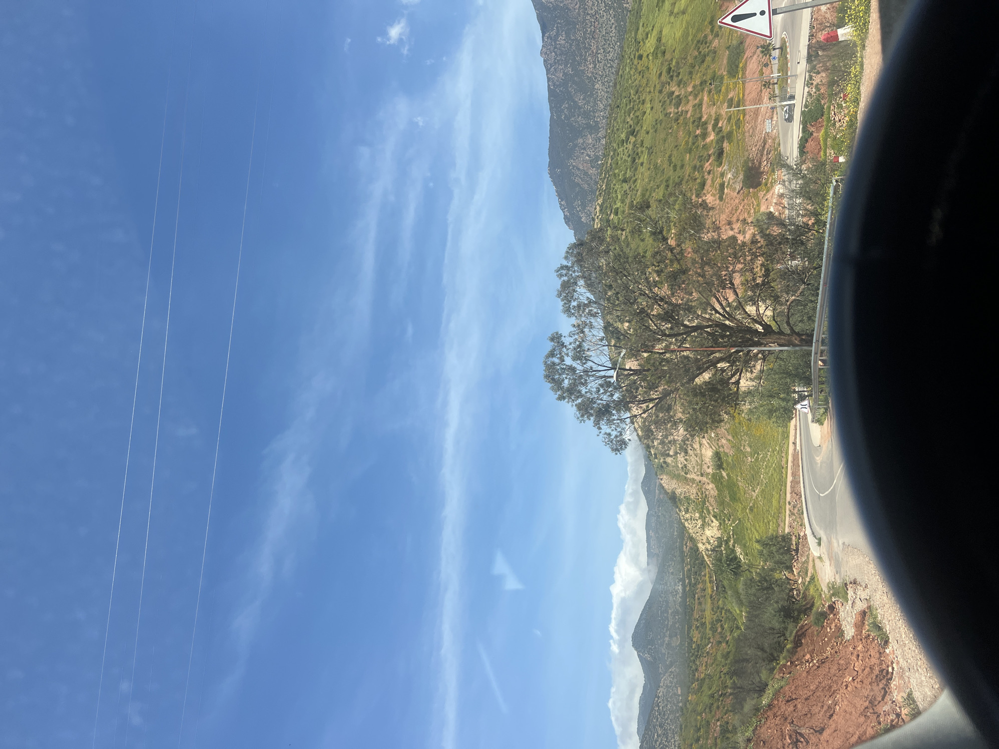 The image shows a landscape in Ouaouizert, Morocco, likely taken from a moving vehicle. In the foreground, a road curves along a hillside with a warning sign visible. The hillside is covered with green vegetation and patches of reddish-brown soil. A tree stands prominently near the road. In the background, mountains rise towards a blue sky with wispy clouds. Power lines are visible in the sky. A portion of the vehicle's interior is visible in the foreground, suggesting the photo was taken through a car window.