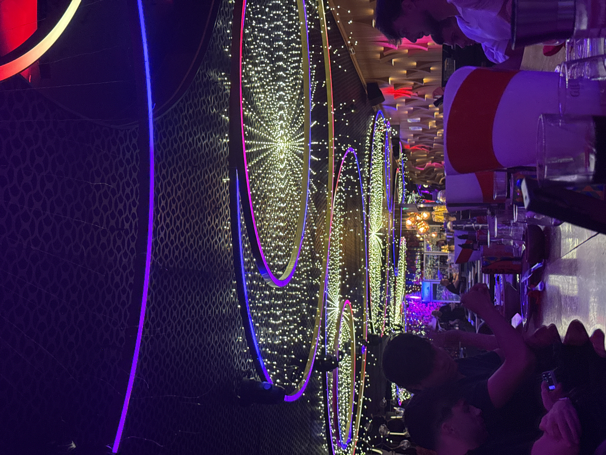 The image depicts the interior of a dimly lit establishment, likely a restaurant or club, characterized by vibrant, multi-colored neon lighting and numerous small, white string lights suspended from the ceiling. Circular light fixtures with purple and blue neon trim are prominent. Several individuals are visible, seated at a counter or table, some with drinks and what appear to be plates. In the foreground, a person in a black shirt and dark headwear is looking down at a phone. To their right, another person in a black shirt is also looking down. Further to the right, a person in a white shirt is visible in profile. The environment features textured dark ceilings and wooden paneling in the background, interspersed with glowing red lights. The overall ambiance suggests a modern, lively entertainment venue. While the image itself does not contain explicit markers, its style and presentation are consistent with venues found in nightlife districts of cities like Sofia, Bulgaria.