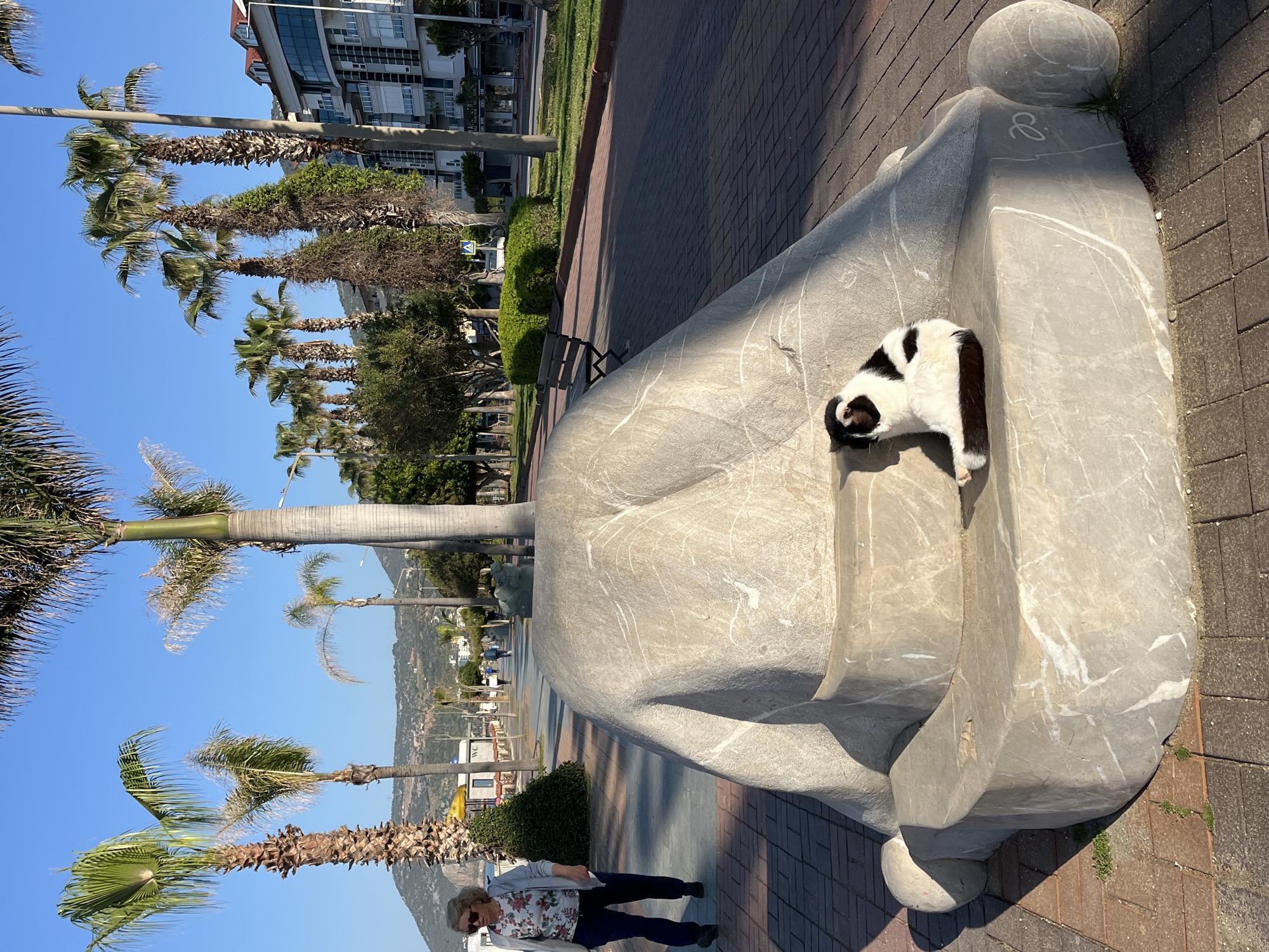 A sunny, daytime scene depicts a seaside promenade in Alanya, Turkey. A middle-aged woman with short blonde hair, wearing a floral top and navy pants, walks near a large, light-grey marble sculpture shaped like a human hand resting horizontally on the ground. A black and white cat is sleeping in the crevice of the palm. Palm trees line the promenade along with green bushes and benches. Buildings are visible in the background under a bright blue sky. The ground is paved with red bricks.
