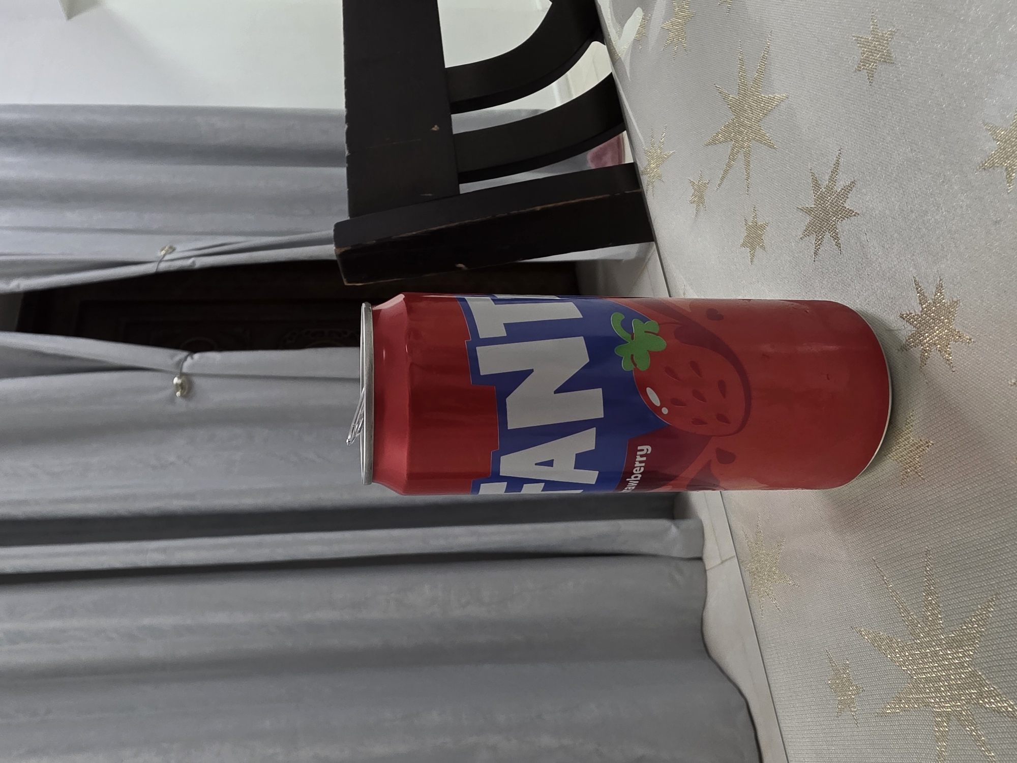 A red Fanta can with a strawberry graphic and "Fanta Strawberry" text is placed on a white tablecloth with gold star patterns. The can is the primary subject of the image. In the background, gray curtains hang. Part of a dark wooden chair is visible on the right side of the image. The scene appears to be indoors, possibly in a dining area or living room. There are no people present. The image does not provide any specific information about the location being in Dibba Province, Oman.