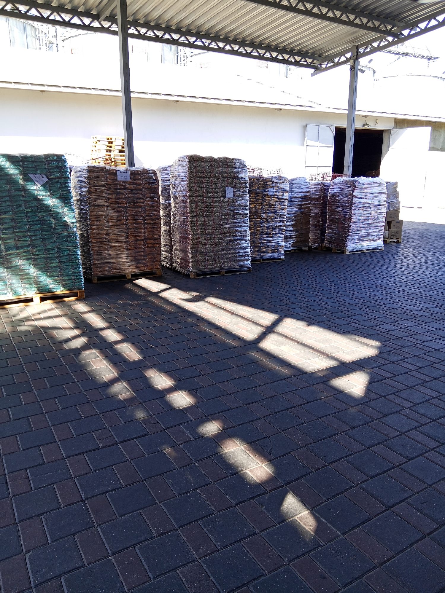 The image depicts an outdoor storage area covered by a metal roof, likely part of a warehouse or agricultural facility in Ceadîr-Lunga, Moldova. The ground is paved with dark gray and reddish-brown interlocking bricks, showing strong diagonal shadows cast by sunlight.
In the background, a white building with a dark doorway is visible, suggesting it is part of the facility. A large gray metal pole supports the roof structure.
The foreground is dominated by stacks of bagged goods, predominantly placed on wooden pallets. The bags are wrapped in clear plastic, revealing their contents which appear to be granular materials, possibly fertilizer, feed, or construction materials. The bags are stacked in rows and vary in color, with one prominent stack on the left appearing in shades of green. Another stack in the center is brown, and further stacks to the right display a multicolored pattern. Some labels are partially visible on the bags, though the text is not legible.
There are no people or animals visible in the image. The scene appears to be a still, organized storage environment, likely a commercial or industrial setting.