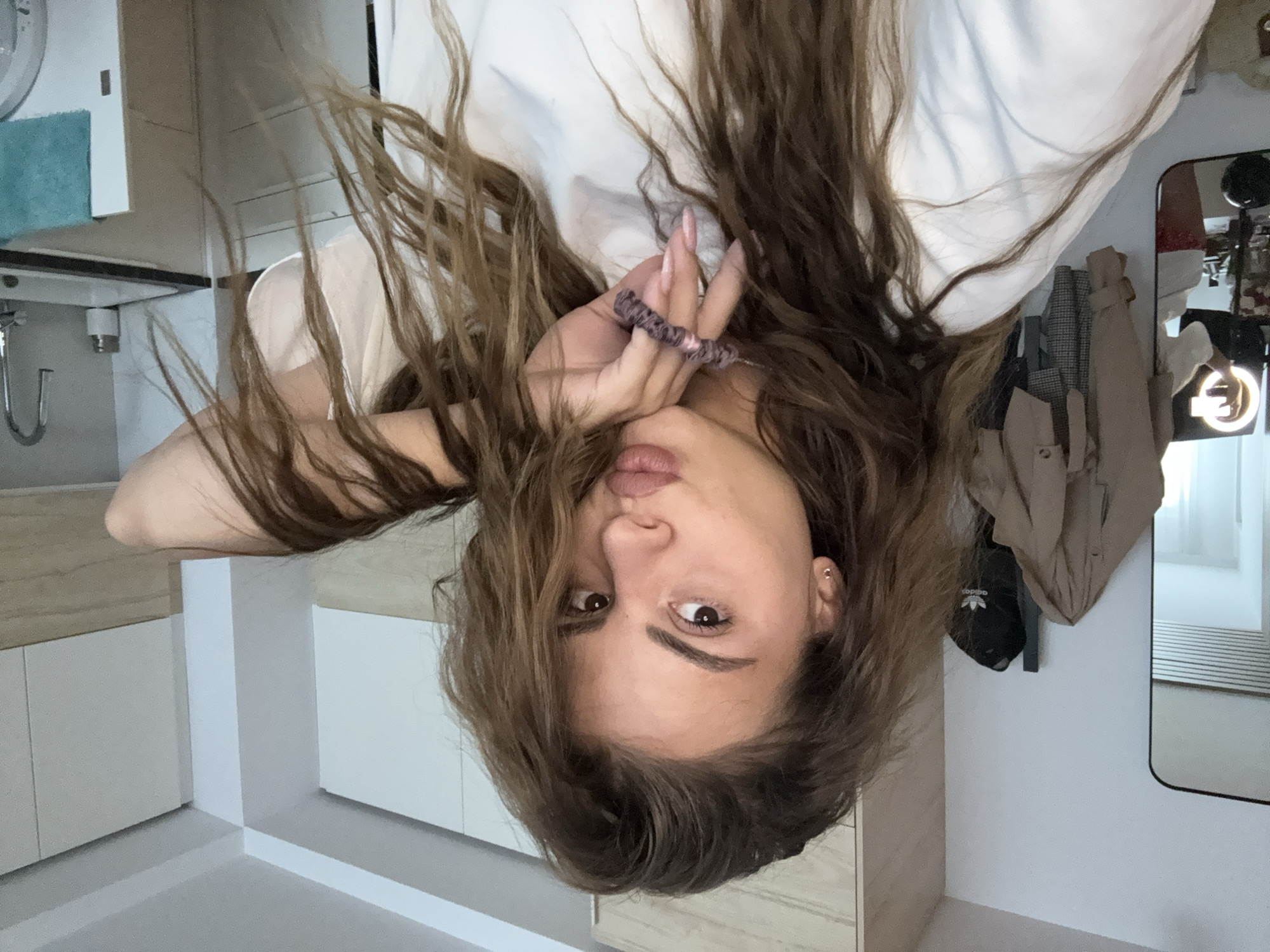 A female with light brown hair is pictured indoors. She is wearing a white top and has a hair tie around her fingers. The background includes a mirror reflecting clothing hanging on a wall, and kitchen cabinets.