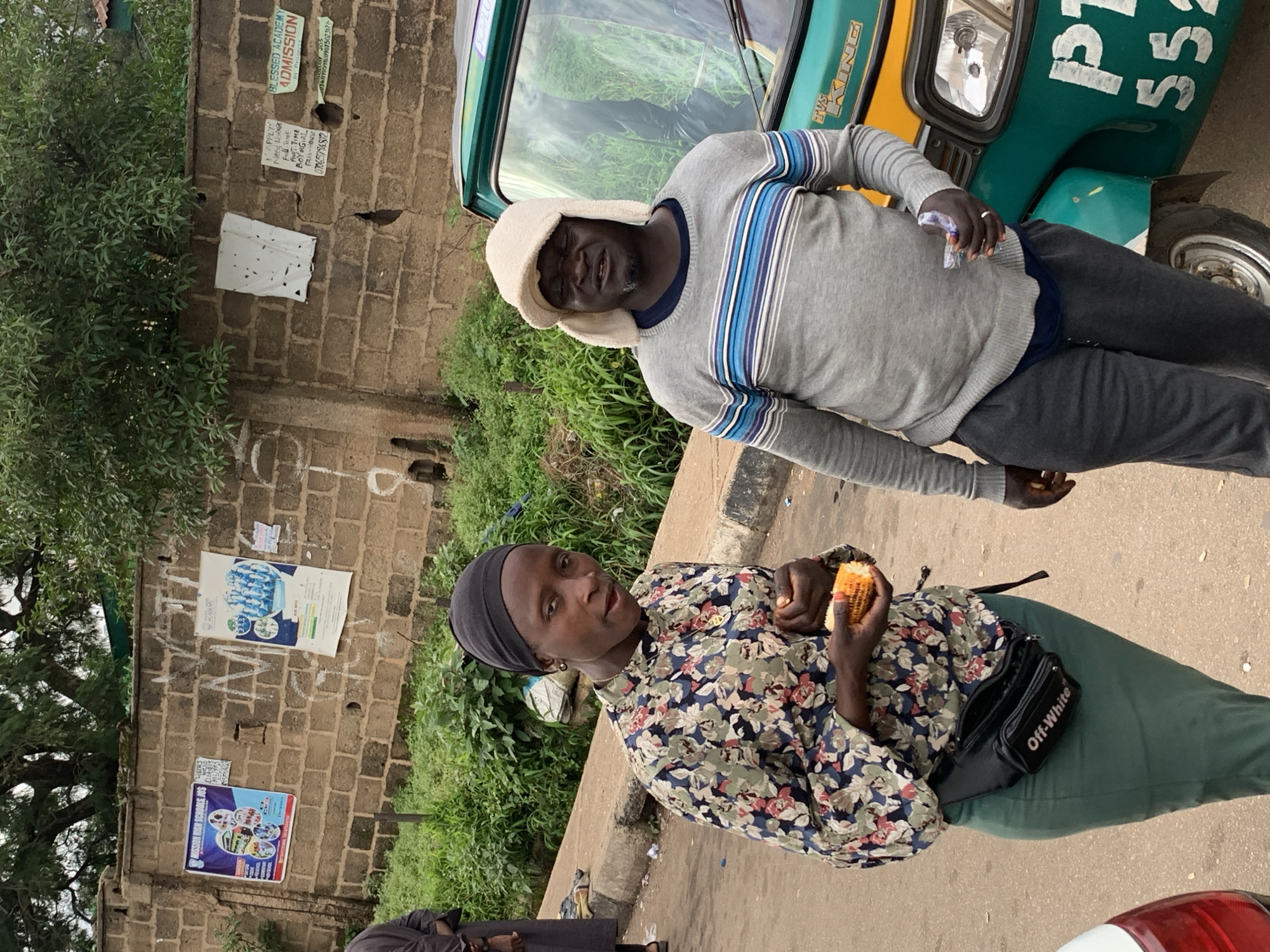 In Barakin Lerawi, Nigeria, a medium shot captures two individuals standing next to a teal and yellow auto-rickshaw on an outdoor street. The woman on the left wears a black headscarf, a floral print shirt with a dark base and colorful flowers, and a dark green skirt. A black fanny pack with "Off-White" branding is worn around her waist. She holds a partially eaten ear of corn in her hands. To her right, a man wears a cream-colored hat with ear flaps, a grey sweater with horizontal blue and white stripes, and dark grey sweatpants. He appears to be holding a small, clear object in his right hand. The auto-rickshaw, a "TVS KING" model, is positioned in the foreground. Behind them, a brick wall is visible, adorned with various posters, including one for "MISSION HIGH SCHOOL JOS" and another for "BLESSED ACADEMY ADMISSION." The environment is lush with green vegetation, and the ground is asphalt. The lighting suggests an overcast or cloudy day.