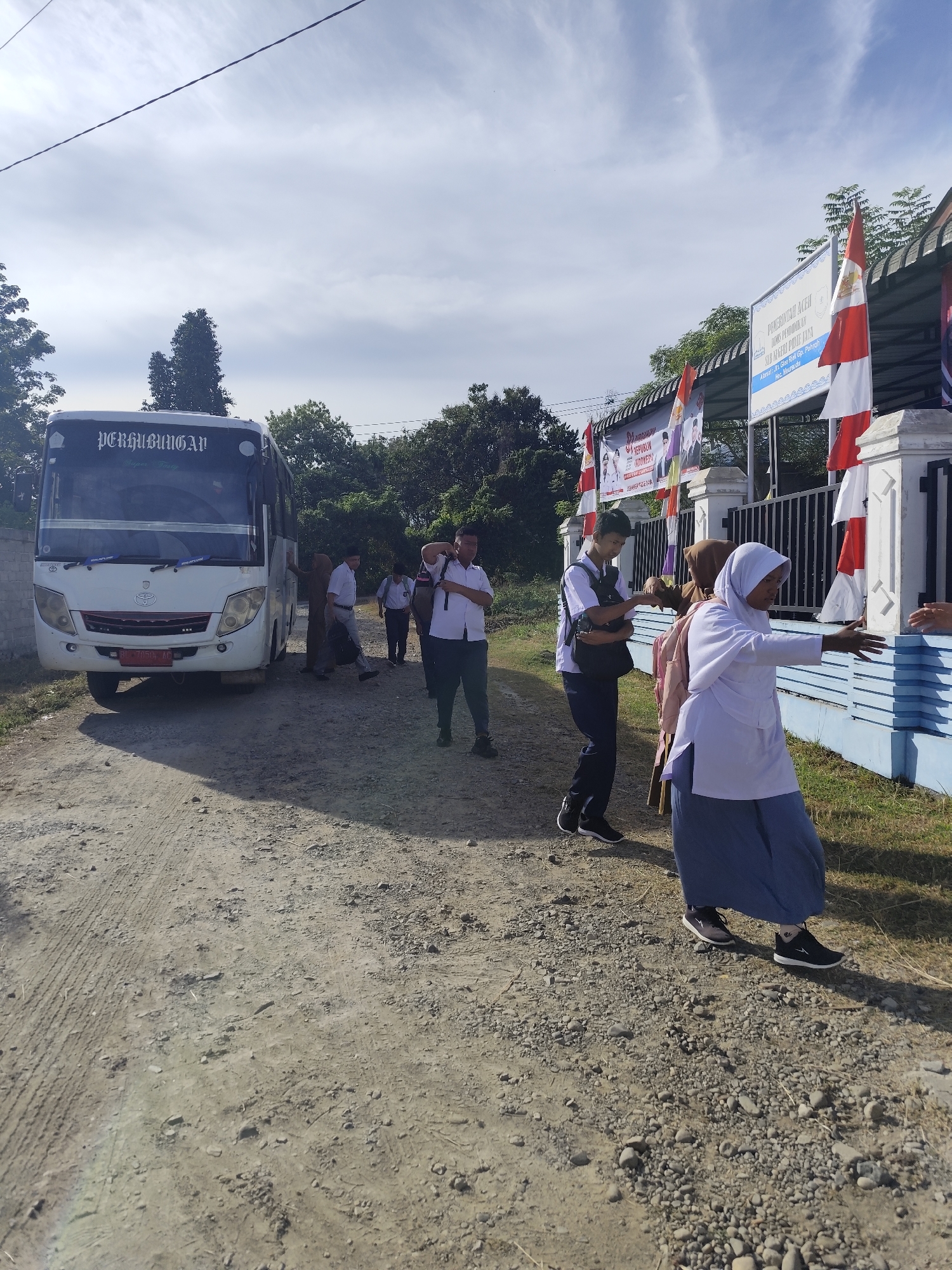 In an outdoor, daytime scene on a dirt road, a white bus with "PERHUBUNGAN" written on its front is parked. Several people are near the bus. On the right side of the image, a building with a white fence and red and white flags is visible. Some of the people are dressed in white shirts, and one person is wearing a hijab and a blue skirt. The environment is rural or semi-rural, with trees and foliage in the background. The ground is uneven and covered with dirt and gravel.