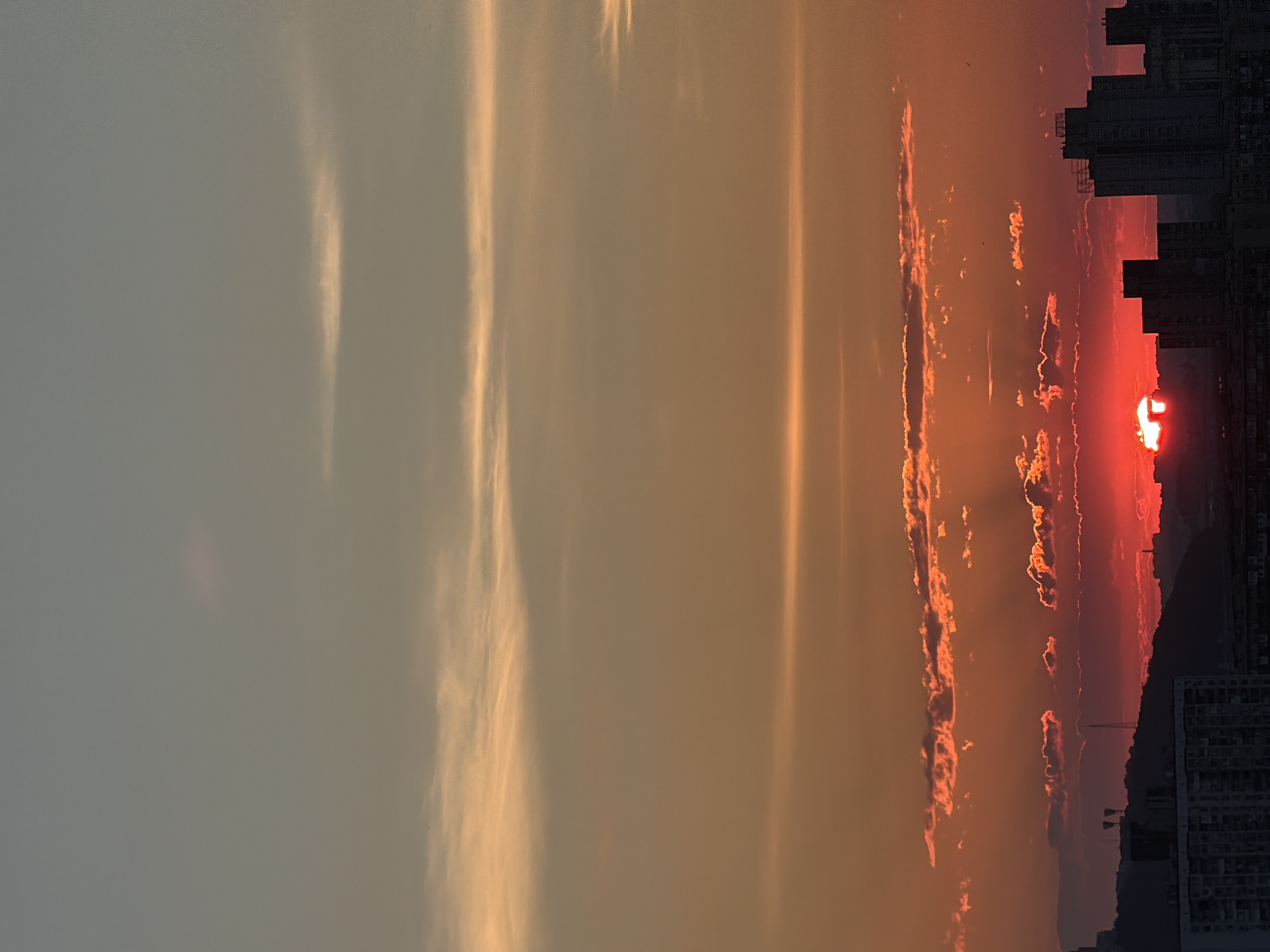 The image captures a dramatic sunset over a cityscape, likely Kyiv, Ukraine, given the provided context. The sun, a fiery orb of intense red and orange, is partially obscured by a line of buildings on the right side of the frame, appearing to set behind them and a silhouetted mountain range. Wispy clouds are illuminated by the setting sun, displaying vibrant shades of pink and orange. These clouds are interspersed with streaks of golden light, suggesting rays of sunlight breaking through.
The sky transitions from a bright, almost white hue on the left, gradually deepening into a warm gradient of orange and red towards the horizon. The left portion of the image is dominated by a pale, muted sky with faint vertical streaks of light, possibly atmospheric effects or reflections. On the right, the dark silhouettes of tall buildings rise against the colorful sky, with one building in the foreground showing detailed window patterns. The overall scene evokes a sense of awe and the beauty of a natural phenomenon. The time of day is clearly sunset, with the warm, saturated colors of the sky being the primary indicator. The weather appears to be clear enough for a vibrant sunset, with the presence of clouds adding texture and drama to the sky. No visible text is present in the image.