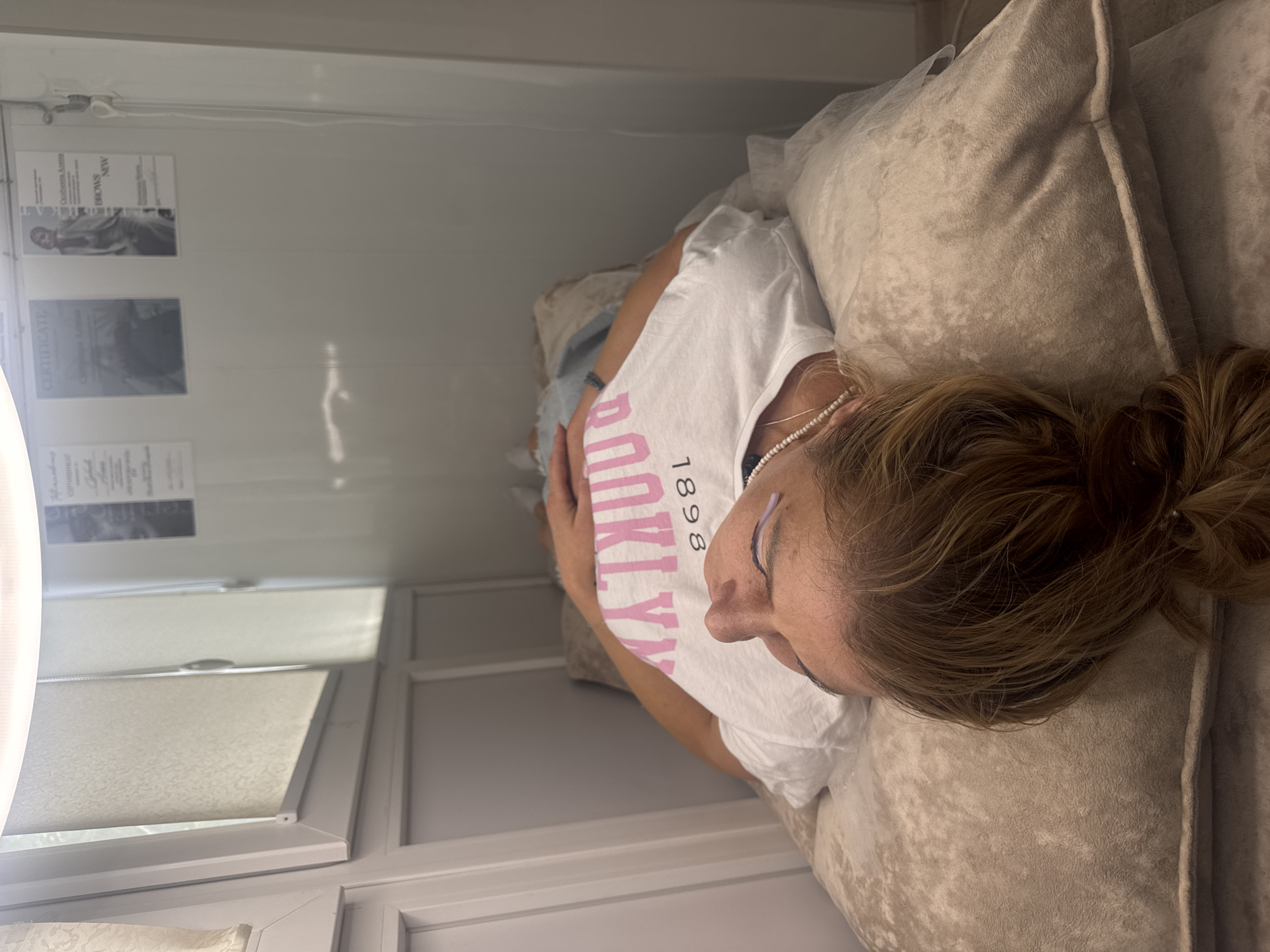 An overhead, medium shot shows a woman lying on her back in a white, narrow room that appears to be a beauty salon. She is wearing a white t-shirt with pink lettering that reads "BROOKLYN 1898". Her eyes are closed, and she has purple tint on her eyelashes. A pearl necklace is draped around her neck. The room has white, paneled walls and a bright light source at the top. There are certificates or posters on the wall, one of which reads "CERTIFICATE". This image was taken in Saratov, Russia.