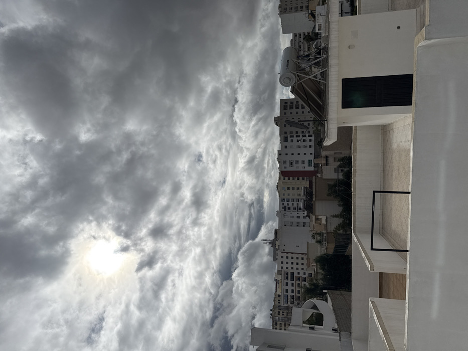 The image presents an elevated view over a residential area within the city of Meknes, Morocco, under an overcast sky. The celestial dome is predominantly covered by dense, grey, and white cumuliform clouds, with the sun appearing as a bright, diffuse light source in the upper-left quadrant, partially obscured by cloud cover.
In the immediate foreground, a flat, white rooftop is visible, featuring a low parapet wall that spans the bottom of the frame. Behind this wall, a lighter-toned tiled terrace is discernible, partially demarcated by a lower white partition, with a dark, rectangular metal frame centrally positioned on it. To the right, on an adjacent rooftop, a small, white, rectangular building with a dark green, vertically paneled door supports a solar water heater system. This system comprises a horizontal, silver-colored cylindrical tank, partially labeled "JUNIPERS," mounted on a dark frame above angled solar collection panels. A gray satellite dish is positioned to the left of the solar water heater.
The midground and background are densely populated with numerous multi-story residential buildings, primarily displaying white or light-colored facades. Some structures exhibit variations, including a building with a vertical red section on its facade and another with a visible antenna or communication tower on its roof. Greenery, consistent with trees or shrubs, is interspersed between some of the buildings. No individuals, specific actions, or interactions are depicted within the scene.