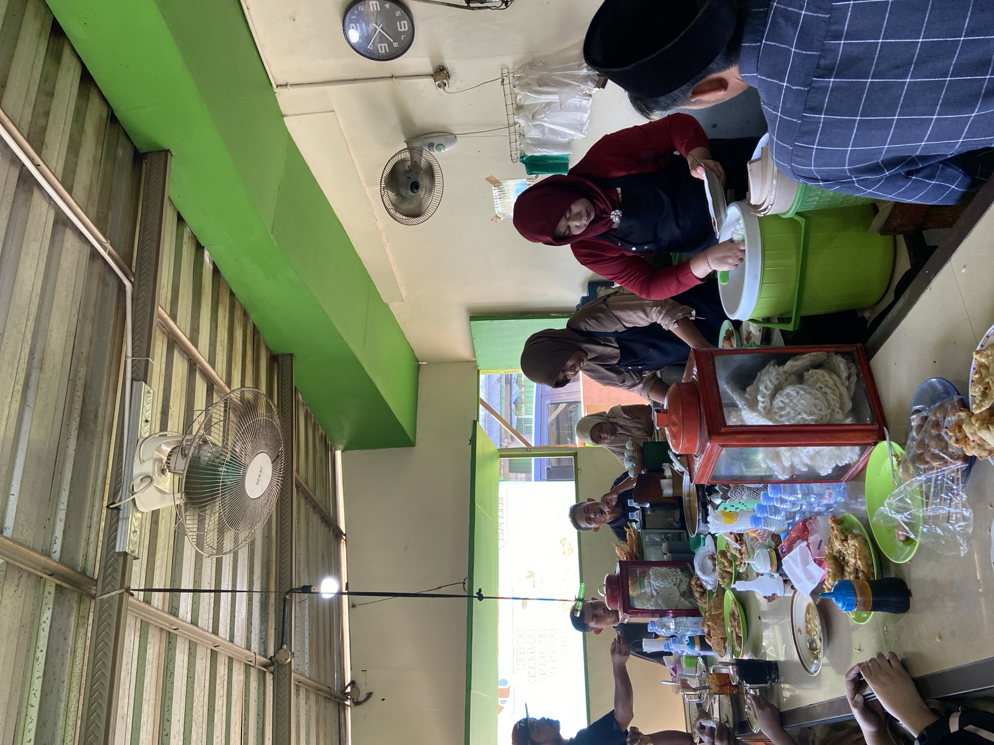 The image depicts a bustling interior scene at a restaurant in Margoyoso, Indonesia. The dining area is open-air with a corrugated metal roof supported by metal beams, painted green. Two large ceiling fans are visible, suggesting a warm climate. The walls are painted cream, with green accents. On the right wall, a round analog clock with black numbers and hands is prominently displayed.
The central focus of the image is a long table laden with food and drinks, where patrons are seated and served. Several people are actively involved in the dining and serving process. Two women, wearing headscarves and black aprons, appear to be serving food. One is scooping rice from a large green container into a plate held by a man. The other woman is tending to food preparation behind the counter.
On the table, various dishes are arranged, including plates of fried chicken, crackers, and bowls of soup. Bottles of water and drinks are also visible, along with condiments. The patrons, a mix of men and women, are engaged in eating and conversation. One man, wearing a black and white checkered shirt and a peci (a brimless cap), is facing away from the camera, interacting with the servers.
The overall atmosphere appears to be that of a casual, local eatery, likely a popular spot for meals in Margoyoso. The scene captures a moment of community and dining, typical of Indonesian culinary culture.