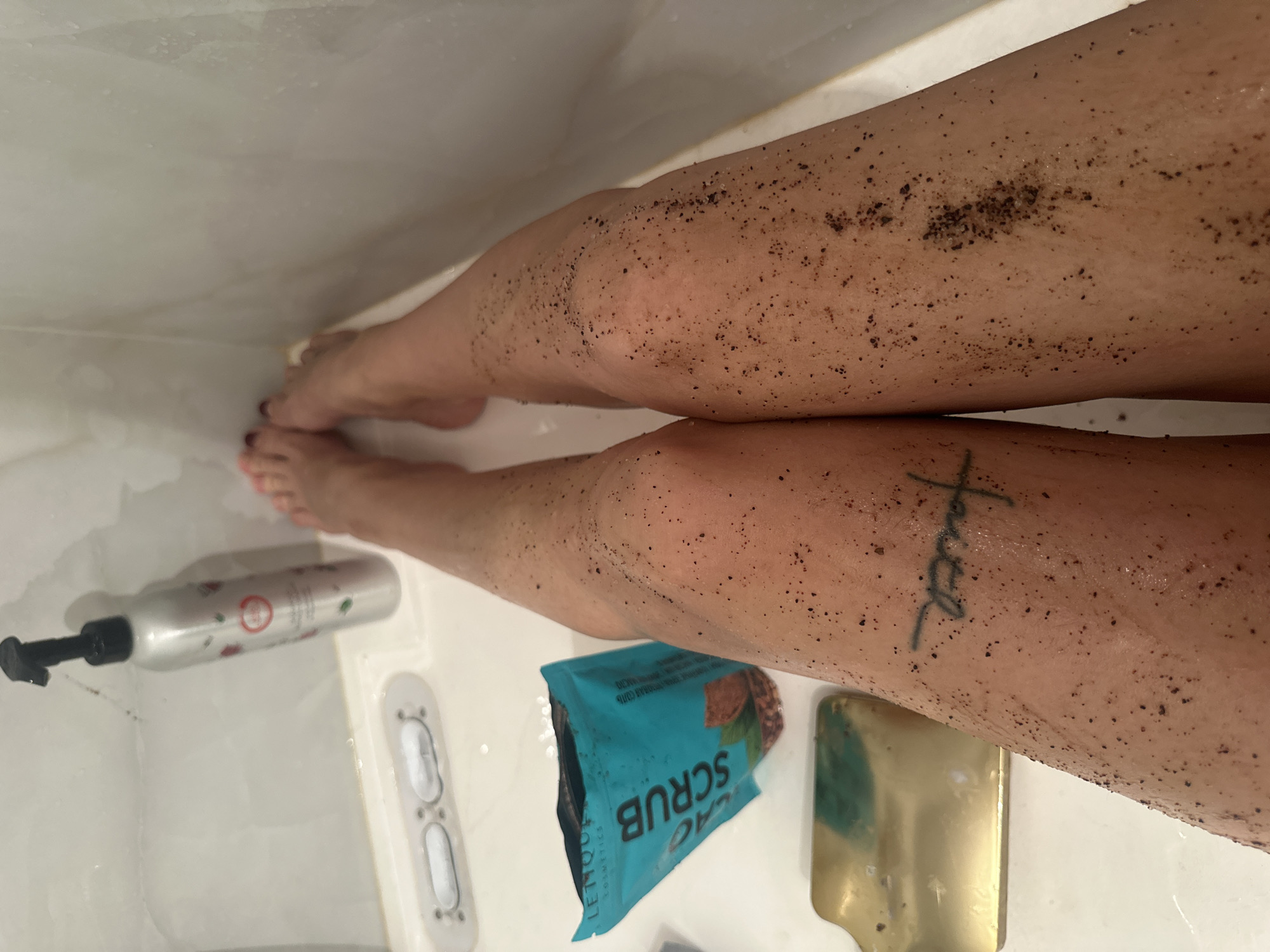 The image presents a top-down view of a person's legs and feet within what appears to be a white shower or bathtub, characterized by wet surfaces. The person's legs are covered in dark, granular particles, suggesting the application of a body scrub. A cursive tattoo spelling "faith" is visible on the inner calf of the right leg, and the toenails are painted with a dark red or purple polish.
On the white shower surface, several objects are visible. A teal-colored packet, partially open, clearly displays the words "CACAO SCRUB" and "LE MOUR COSMETICS," confirming the nature of the scrub being used. Nearby, a rectangular, reflective golden object, possibly a personal care tool, rests on the surface. To the left, a silver pump bottle with a black dispenser head lies horizontally, featuring a white label with subtle red and green design elements. A white panel with two oval controls or indicators is also present in the shower basin.
The activity depicted is a personal care routine, specifically the use of a body scrub for exfoliation. The scene is set in a bathroom. No specific time of day, weather conditions, or emotional states are discernible from the image. There are no visual cues within the image to indicate the provided location context of Gavrilkovo, Russia.