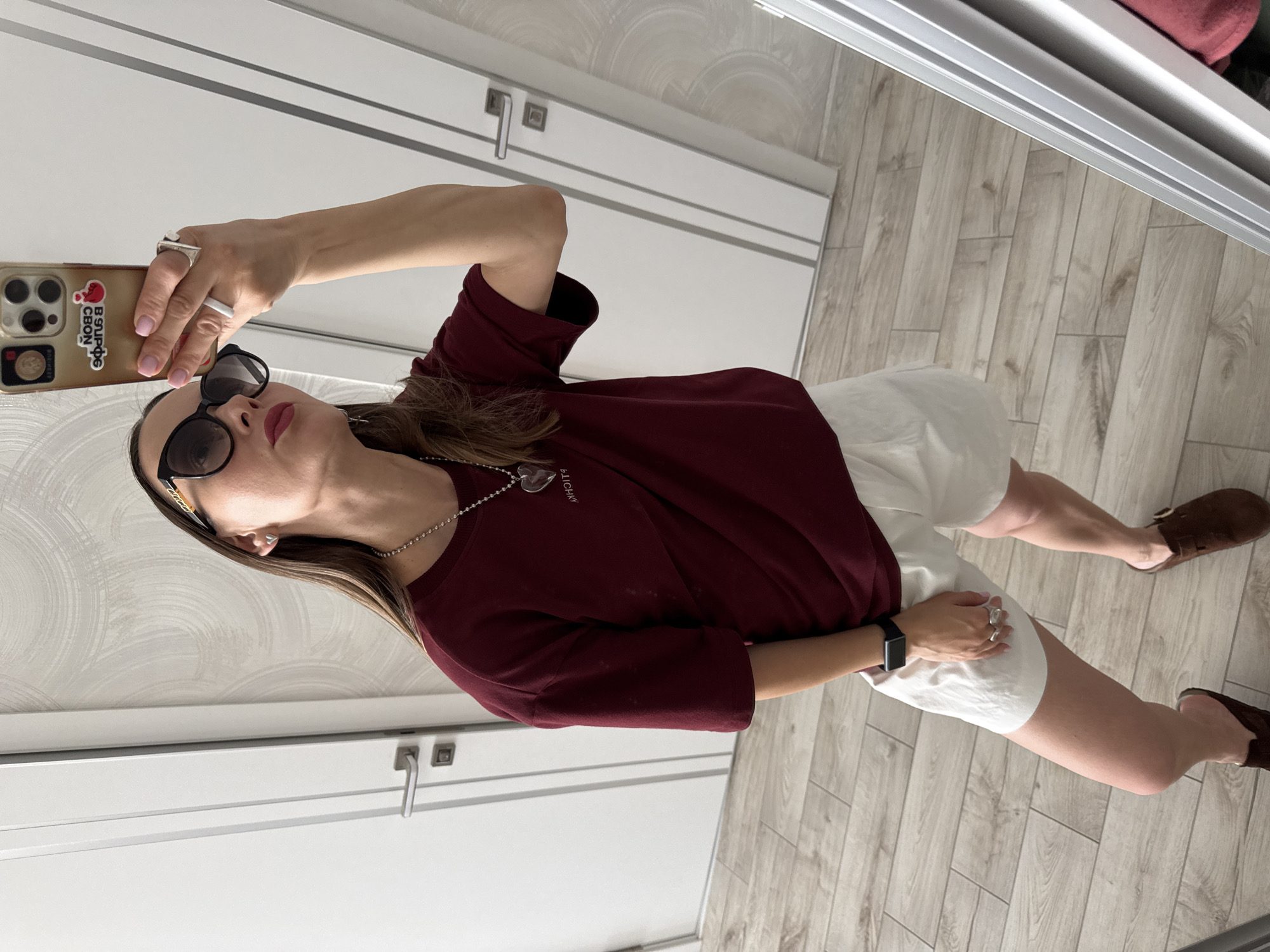 A young woman is taking a mirror selfie in what appears to be a home or hotel room. She is wearing a maroon t-shirt, white shorts, sunglasses, and brown clogs. She has on a necklace with a heart-shaped pendant and several rings. Her phone, which is gold with stickers on the back, is held up to take the picture. The background shows doors with white paneling and a light wood-grain floor. The scene is well-lit and the woman is the clear subject of the photograph. The location is not explicitly indicated as Sochi, Russia, but the visual style of the doors and interior are consistent with some modern residential or hotel interiors found in many urban areas.
