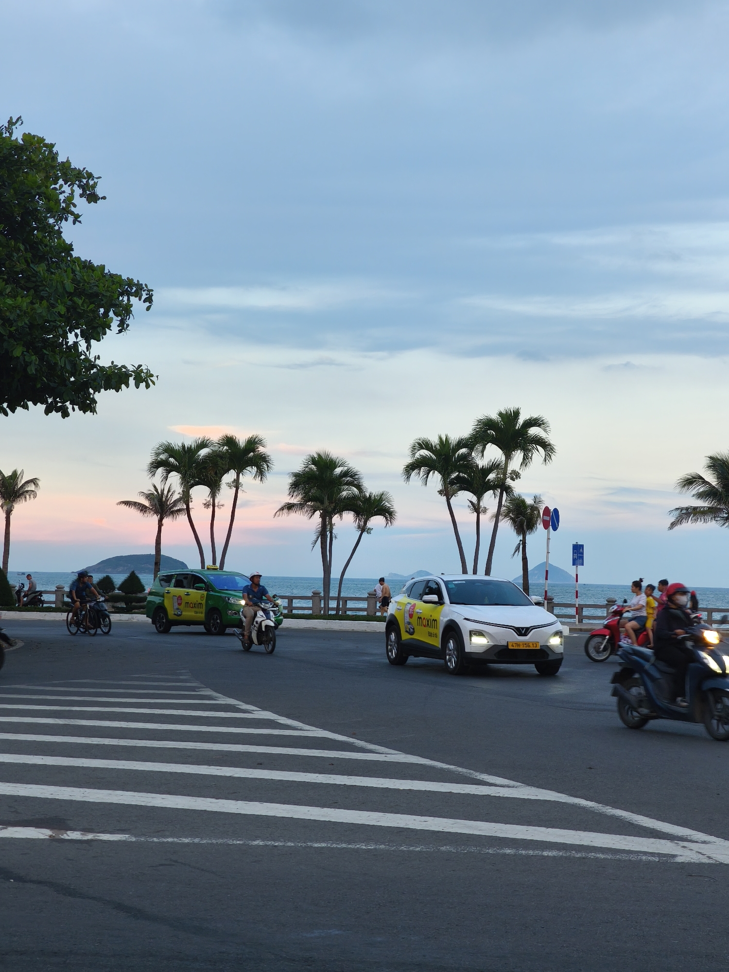 The image shows a street scene in Bac Nha Trang Ward, Vietnam, during twilight. The sky is a mix of blue and pink hues, with some clouds visible. Palm trees line the street, with the ocean and a distant island in the background. The road has a crosswalk in the foreground. Several vehicles are present, including a green Maxim taxi, a white Maxim taxi, a motorbike carrying two people, and another motorbike carrying one person. There are also a few pedestrians visible along the sidewalk. The overall atmosphere is serene and suggests a coastal city at dusk.