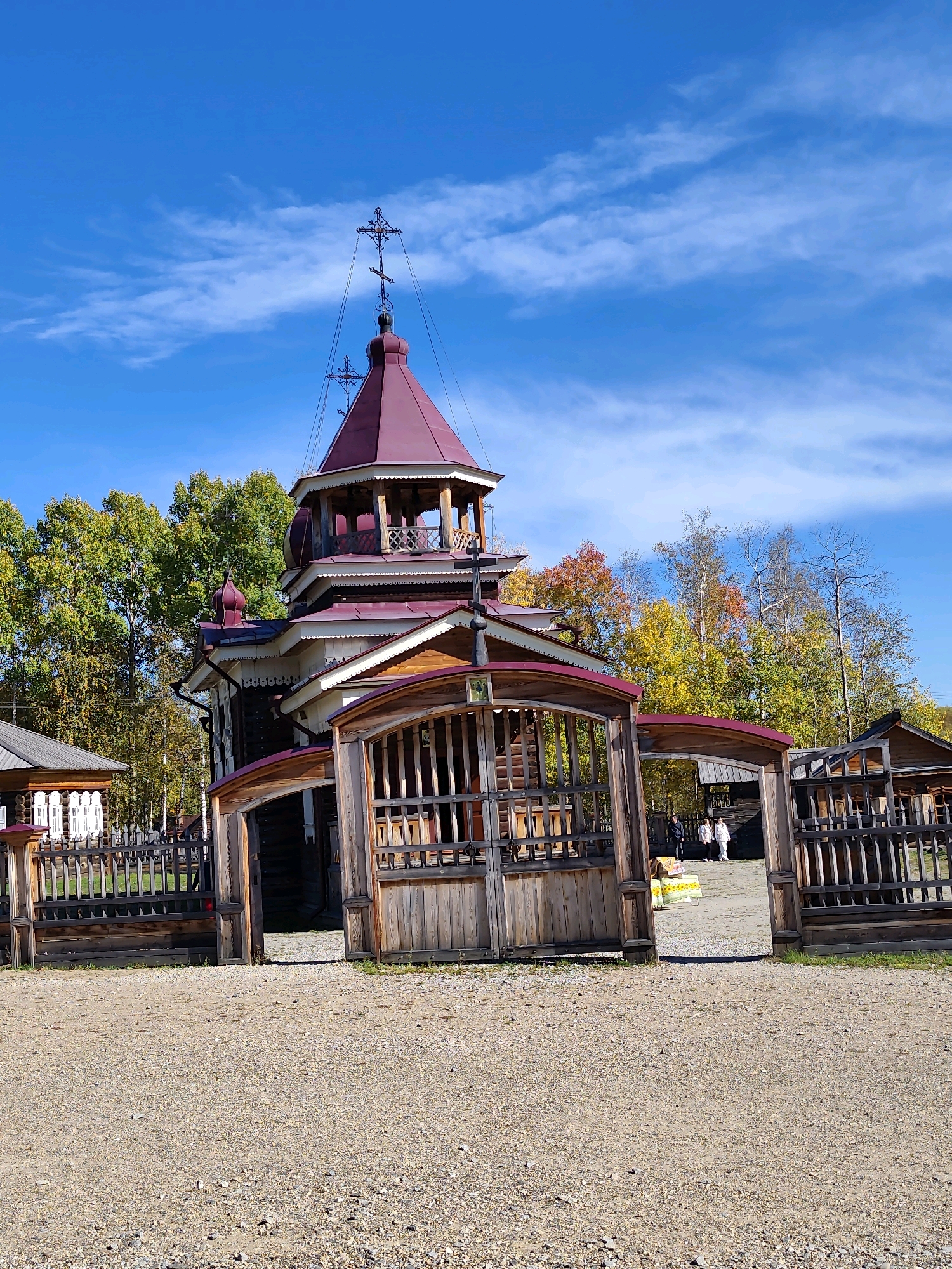 The image shows the entrance to an outdoor museum in the city of Taltsy, Irkutsk Oblast, Russia. A wooden church with a red roof and a bell tower is visible in the center of the frame. The church is adorned with crosses on its rooftops. A wooden gate and fence surround the church grounds. The surrounding environment features a gravel path leading to the church and a backdrop of autumn trees with colorful foliage. Several people are visible in the distance, entering the church grounds through the gate. The sky is a clear blue with scattered clouds, indicating a pleasant, sunny day.
