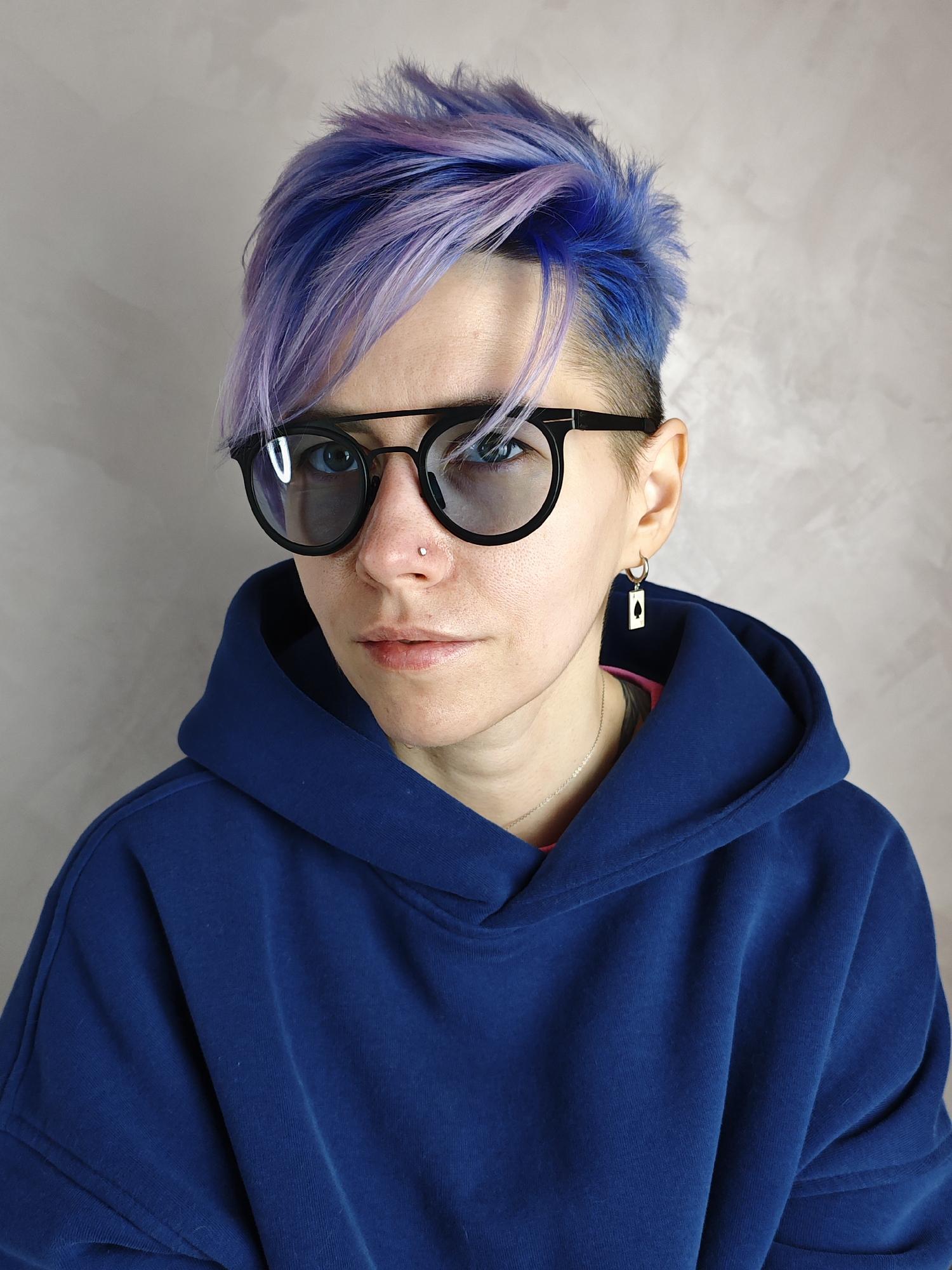 A person with short, blue and purple hair is wearing black round-framed glasses and a blue hoodie. They have a nose piercing and are wearing a rectangular earring with a black spade symbol. The background is a mottled grey wall. The image was not taken in Izhevsk, Russia, as there is no visual information to confirm this.