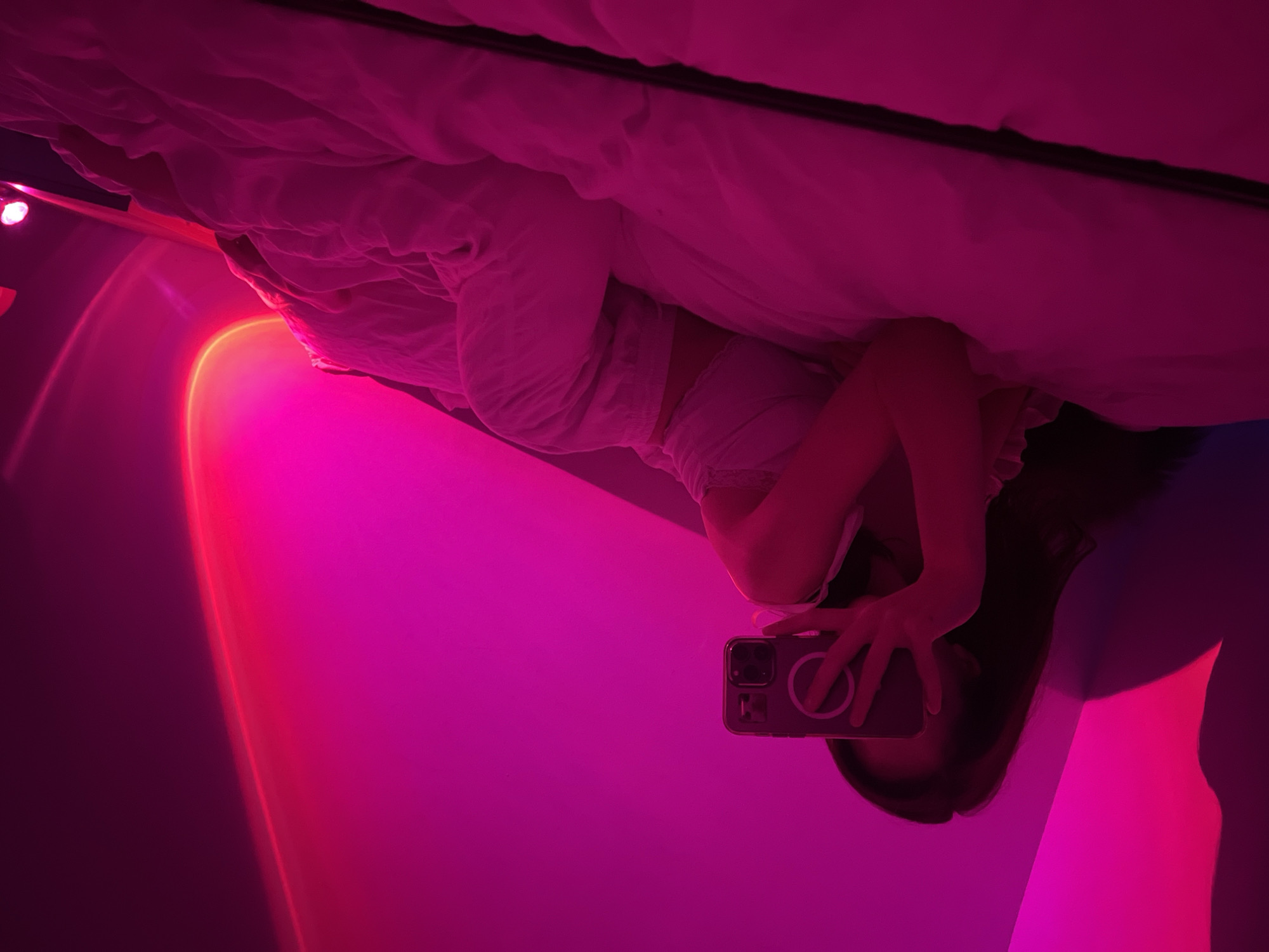 A person is lying on a bed, holding a smartphone to take a mirror selfie. The room is bathed in pink and purple light, likely from a sunset lamp. The person is wearing light-colored sleepwear. The bed appears to have white linens. The background is dominated by the gradient of the ambient light. The overall mood is relaxed and intimate. The image does not provide specific clues about the time of day beyond the colored lighting, nor does it offer weather or specific location cues beyond the user-provided context of Ternopil, Ukraine. No visible text is present in the image.