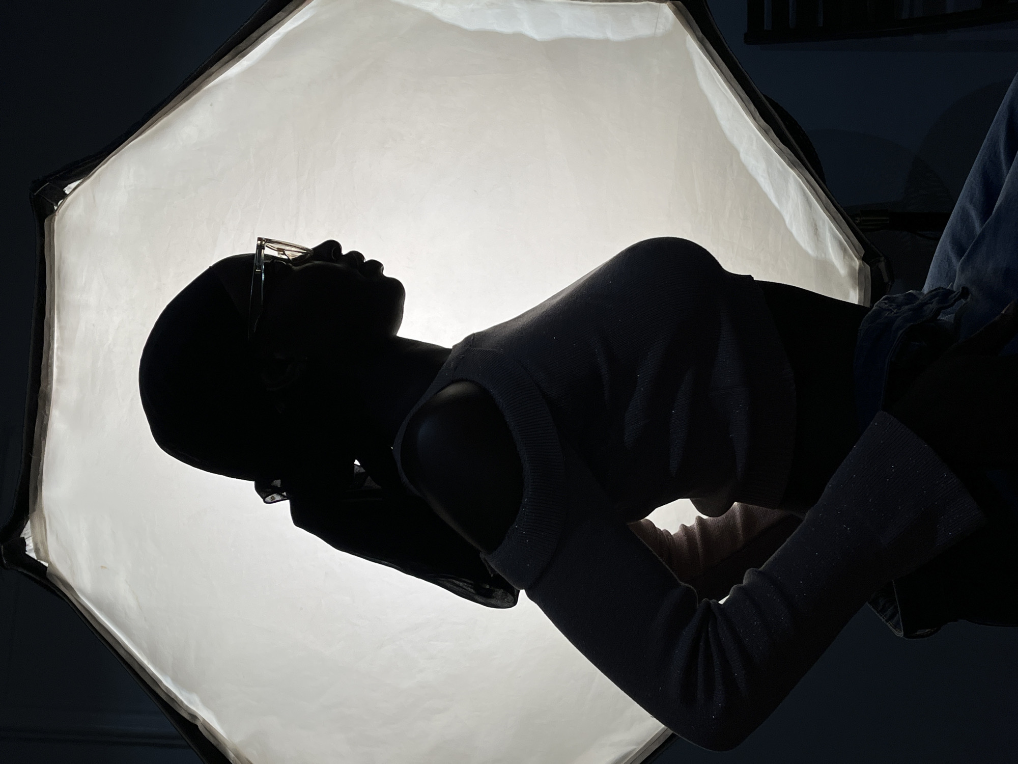 The image shows a person with dark skin, likely female, wearing glasses, with their head in the lower left quadrant of the image, angled towards the upper right corner. Their silhouette is stark against a bright, white, octagonal softbox. The person is wearing a light-colored sweater with long sleeves. The background is dark, but hints of an indoor setting can be seen, including what appears to be a window frame or shelving in the upper right quadrant. The image does not provide information to conclude the location is Barakin Akawo, Nigeria.