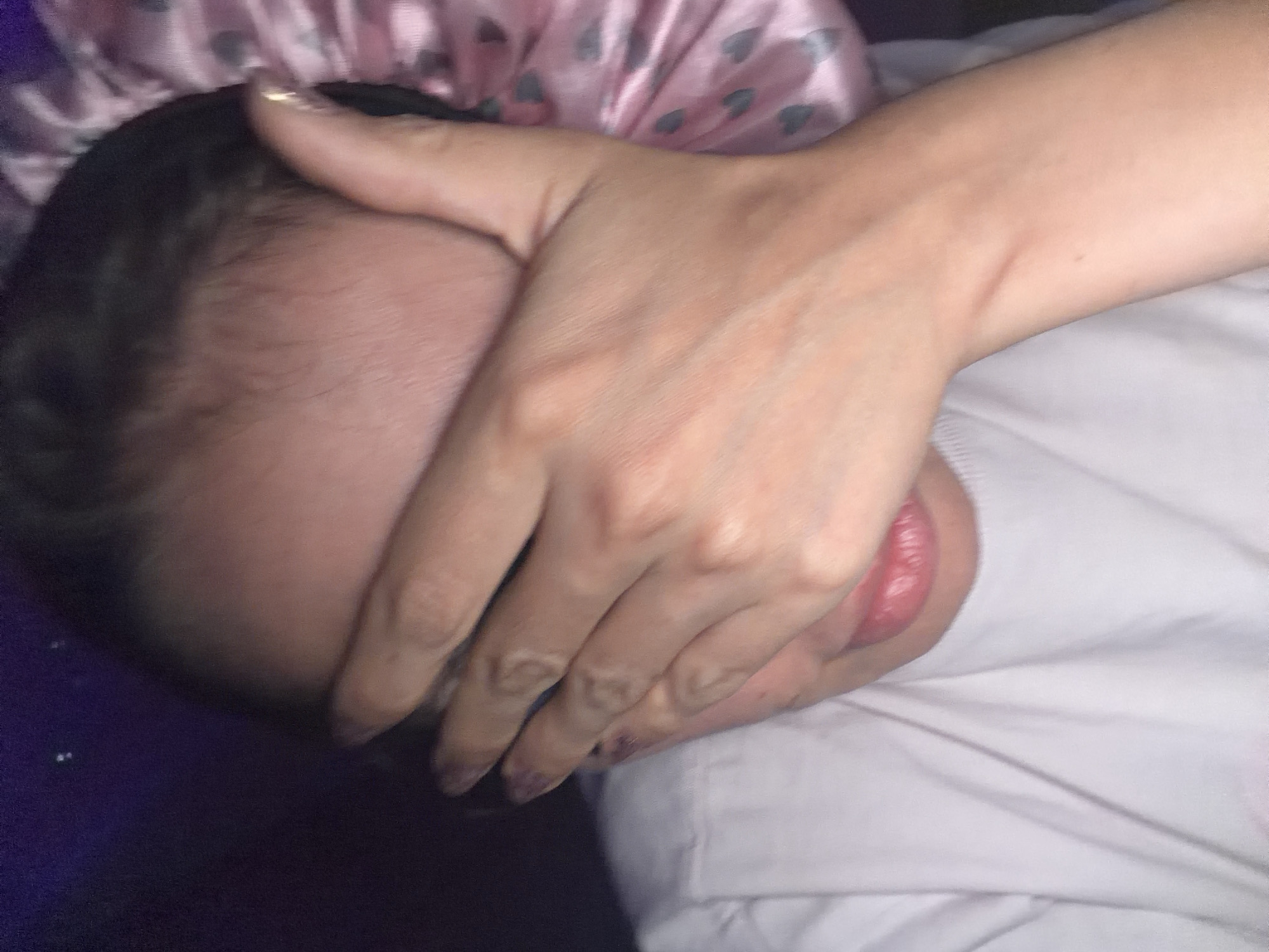 The image presents a close-up, dimly lit indoor scene featuring an individual, located in Tersefanou, Cyprus. The subject's face is largely obscured by their right hand, which covers the eyes and upper portion of the nose. The lower part of the face, including the mouth with full, pink lips slightly parted, is visible beneath the hand. The subject is wearing a white crew-neck t-shirt, showing mild creasing across the chest. Dark hair is visible along the hairline on the right side of the forehead, beneath a pink head covering patterned with small, darker grey hearts, visible on the upper right of the frame. The fingernails on the visible right hand appear manicured, with the nail on the index finger exhibiting a metallic gold color or a distinct gold ring. The background is predominantly dark purple, with two minute, indistinct light reflections present in the upper left quadrant. A segment of the subject's left arm or elbow is visible on the lower right, resting on an unseen surface.