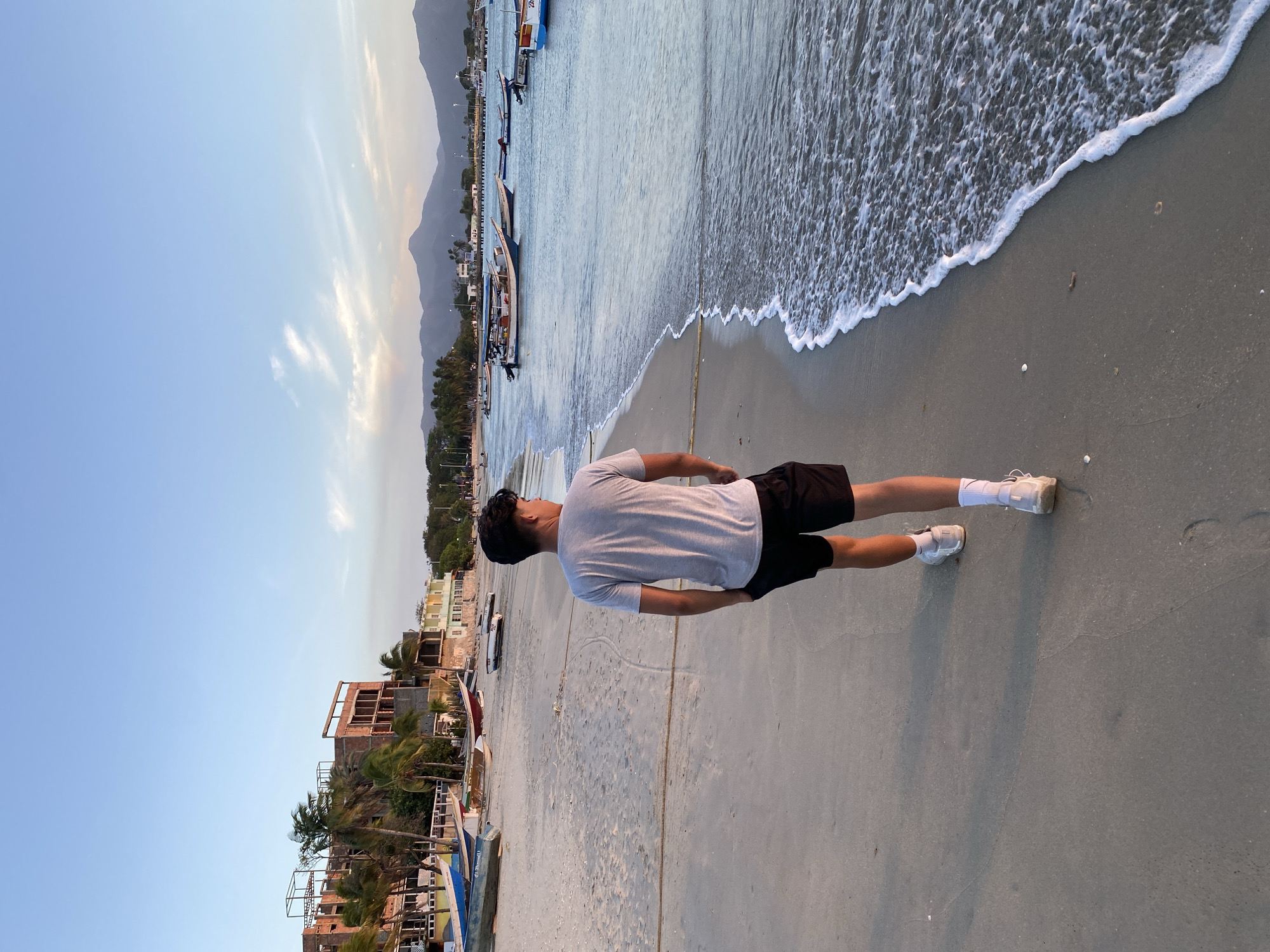 A medium, horizontally oriented shot depicts a man standing on a beach, looking toward the ocean. He is wearing a light gray t-shirt, black shorts, white socks, and white sneakers. The ocean is on the right, with visible waves. Boats line the beach to the left of the man. In the background, there are buildings and mountains. The sky is blue with light clouds. The scene is possibly near the city of Juan Griego, Venezuela, based on the buildings and landscape.