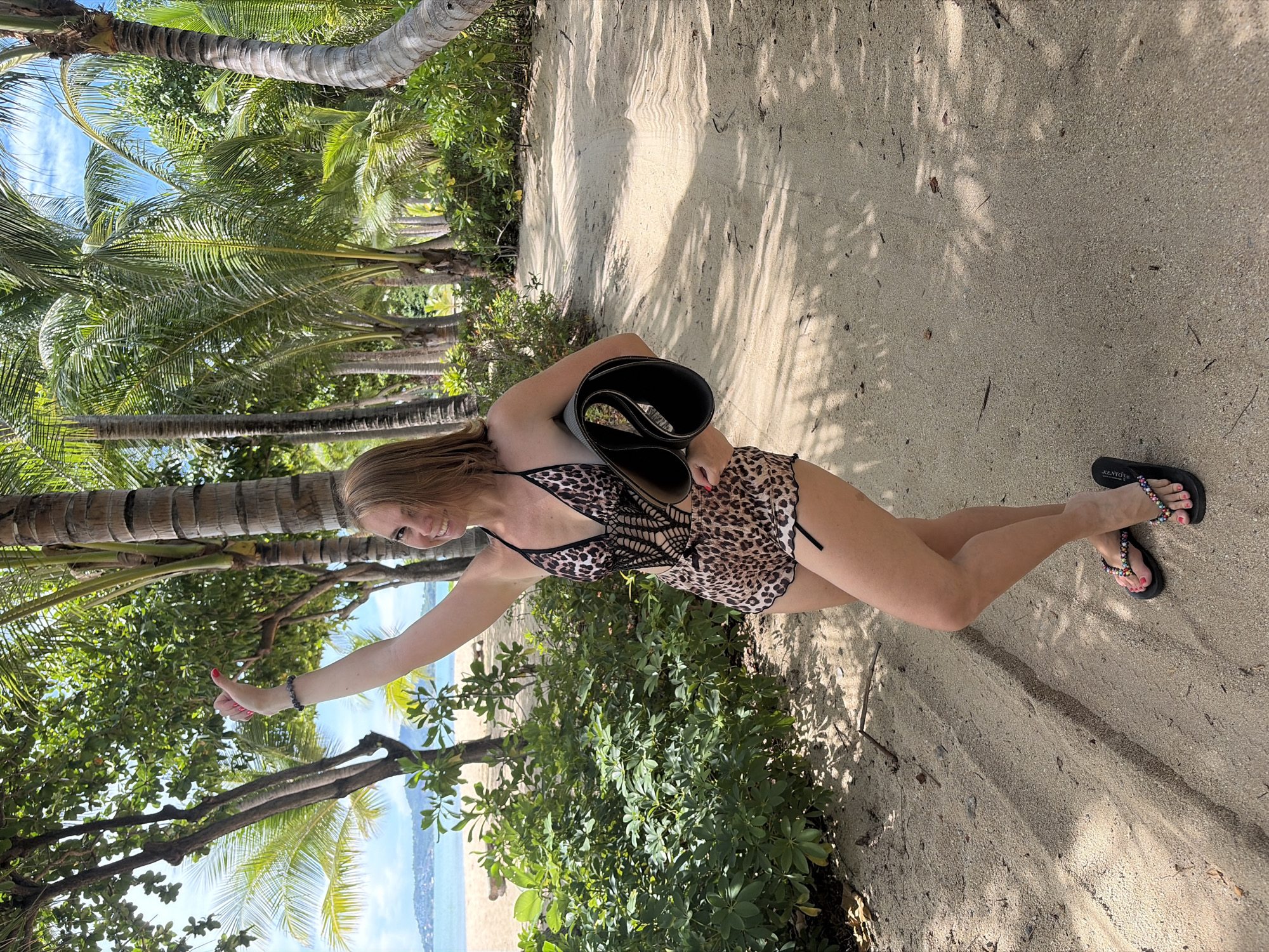 A woman with blonde hair and a leopard-print swimsuit is standing on a sandy path lined with palm trees. She is holding a rolled-up yoga mat in her left arm and waving with her right hand. She is wearing black flip-flops adorned with colorful beads. Behind her, a beach and the ocean are visible under a bright, cloudy sky. The path appears to be in a tropical location, possibly near Baan Bo Phut, Thailand.