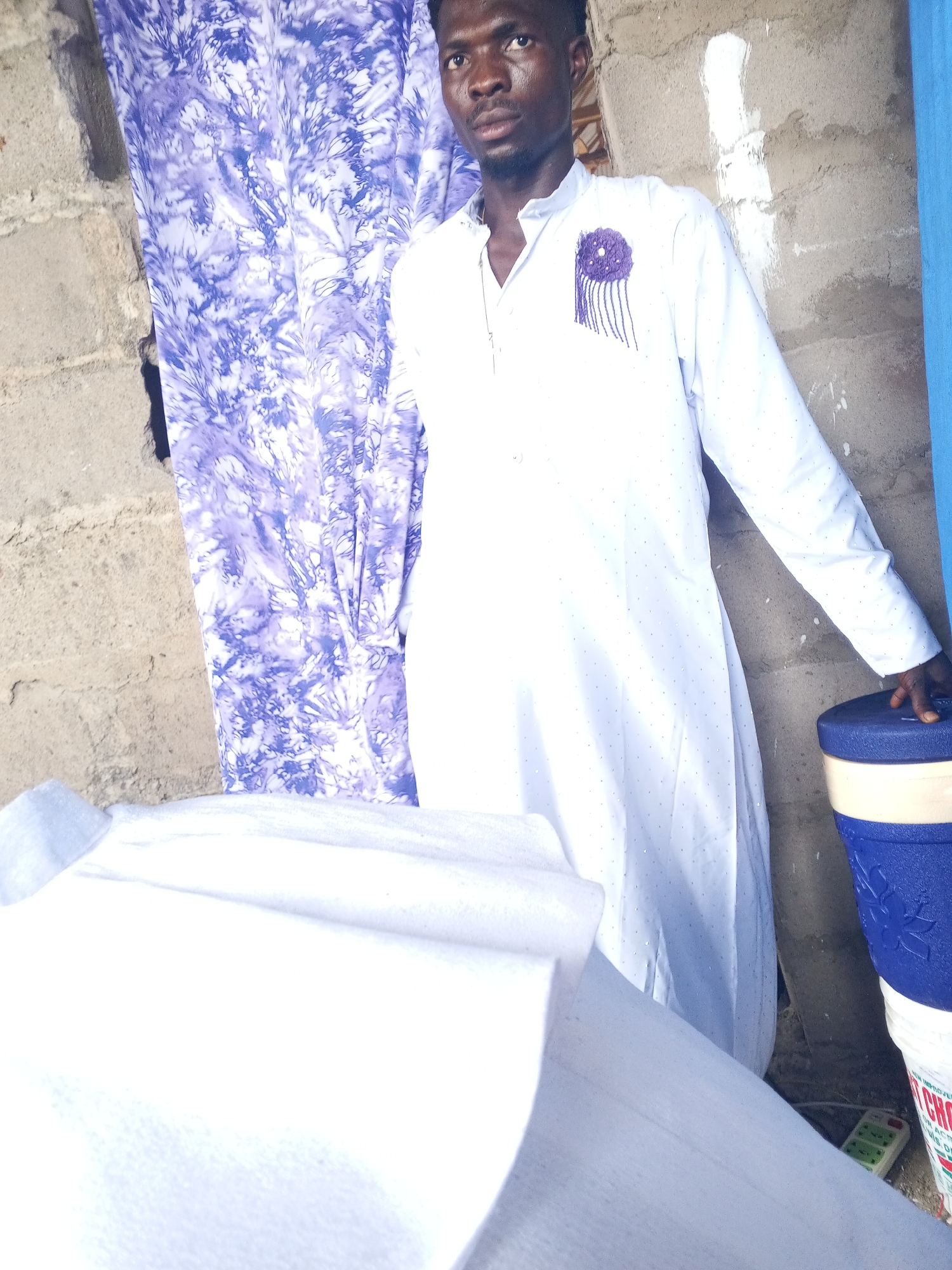 A man in a white outfit stands partially obscured by white fabric, near a building. He has short dark hair, and his garment features purple embroidery. Behind him, a purple and white patterned cloth hangs against a concrete wall. The building itself appears to be constructed from rough concrete blocks, some bearing patches of white. Adjacent to the man, to the right, is a blue and white container. In the foreground, some white fabric is positioned. Also in the foreground there is what appears to be an extension cord.