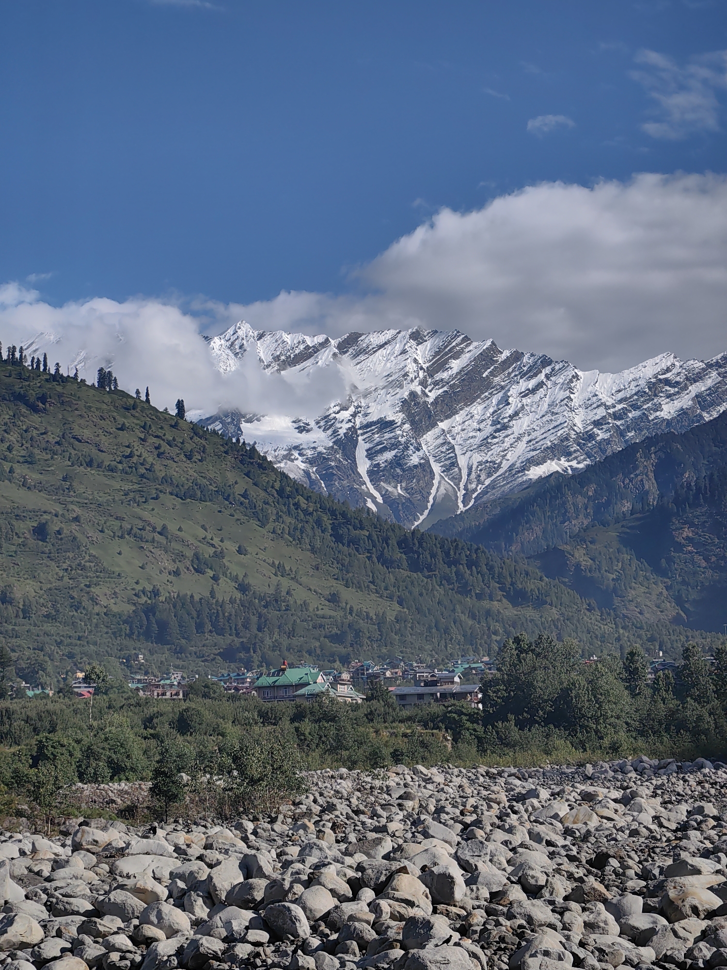 The image captures a majestic mountain landscape with a clear blue sky partially obscured by white clouds. Snow-capped peaks dominate the background, their rugged slopes etched with shadows and streaks of white snow. In the middle ground, a verdant green hillside slopes down towards a cluster of buildings that form part of the village of Bahang, India. These buildings feature a mix of architectural styles, with several houses showcasing traditional pitched roofs, some of which are green. The foreground is a dry riverbed filled with numerous large, smooth, grey stones. The scene is bathed in natural sunlight, creating a serene and picturesque atmosphere.