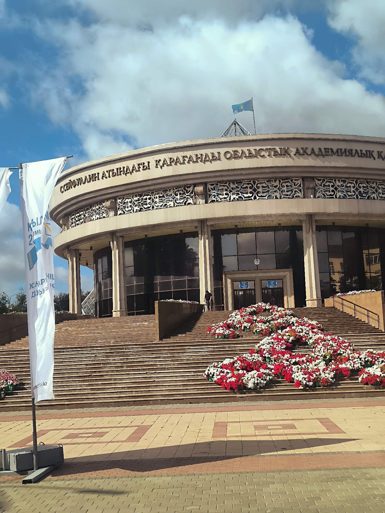 The image depicts the front of a large, circular building with an inscription in Cyrillic script across its facade. The building appears to be a public institution, possibly a government or cultural center, given the flag flying from the roof and the formal architectural style. In front of the building, a wide set of stairs ascends to the entrance. The stairs are adorned with numerous floral arrangements, predominantly in red and white, creating a festive or commemorative display.
Two tall white banners stand on the left side of the image, featuring text in Cyrillic and possibly other languages. One banner has graphics that suggest a map or educational symbol. The foreground is a paved area with a patterned surface, and shadows suggest it is daytime. The sky above is partly cloudy. A person is visible on the stairs, near the entrance, possibly an employee or attendant. The overall impression is one of a significant civic building in a well-maintained urban setting.
