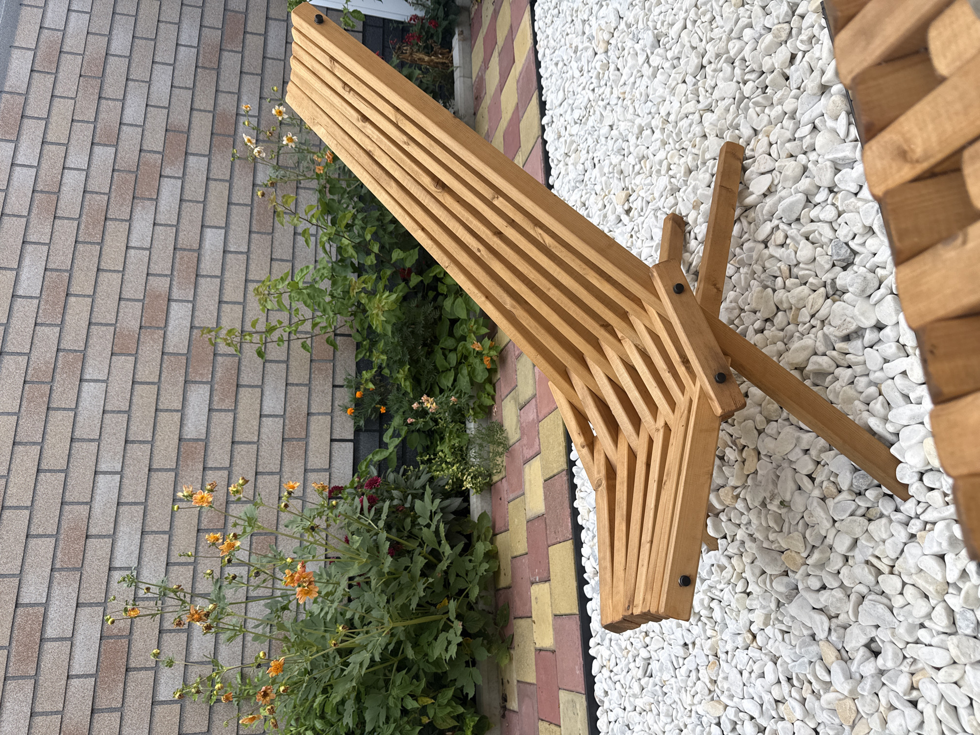 A wooden lounge chair is situated on a bed of white pebbles in front of a brick wall. Green plants with orange flowers are planted between the pebbled area and the wall. The ground in front of the plants is paved with colored rectangular stones.
