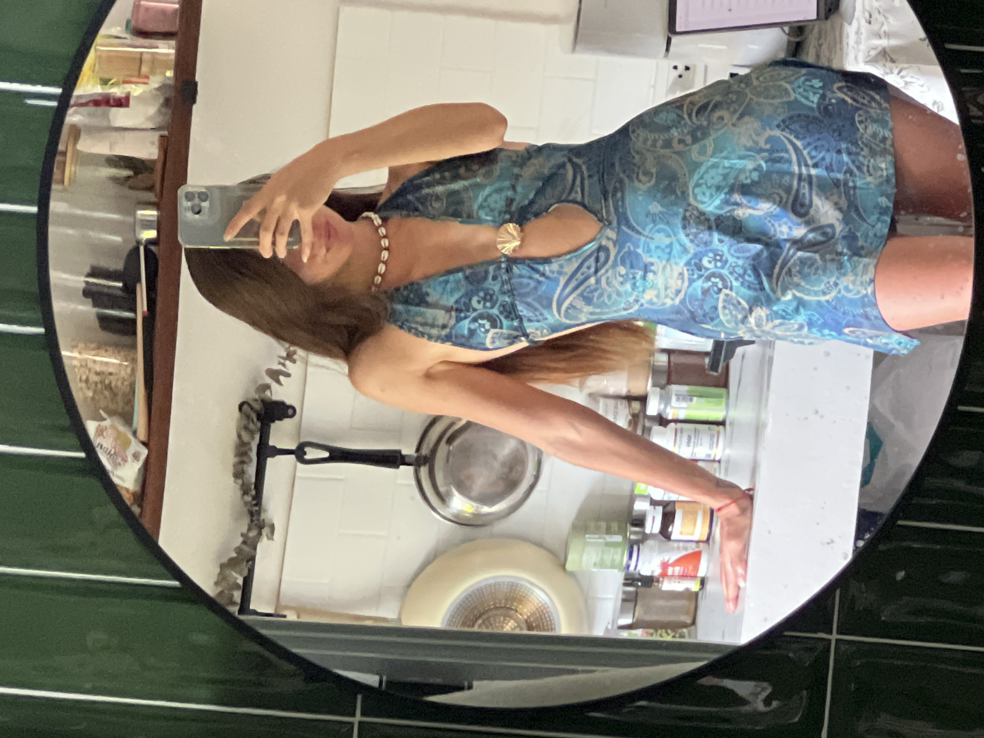 A woman stands in what appears to be a kitchen, taking a mirror selfie. She is wearing a blue, patterned halter dress with a deep V-neckline and a seashell-like embellishment in the center. Around her neck, she wears a seashell choker. Her arm is resting on a countertop with various bottles and jars of supplements. In the background, pots and pans hang on the wall, and shelves are stocked with kitchen items. The lighting suggests it is daytime, possibly indoors with natural light. The environment is domestic, with a focus on personal presentation and everyday objects.