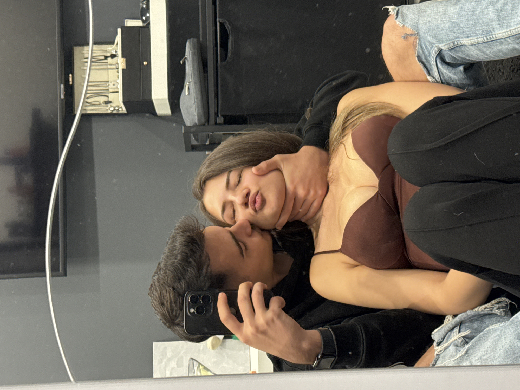 This mirror selfie, taken indoors, captures a young couple in an intimate and playful moment. The image is oriented vertically, showing their reflection as if they are lying down or leaning back.
On the left, a young man with dark hair is holding a black smartphone with multiple cameras (likely an iPhone) to take the picture. He wears a dark hooded sweatshirt and a black watch on his left wrist. His face is turned towards the woman, eyes closed, seemingly kissing her cheek or neck. His hand is playfully placed around her neck or jaw. Part of his ripped blue jeans are visible on the far right.
The young woman is embraced by the man, making a playful expression with her eyes closed and lips puckered. She has brown and blonde hair and is wearing a dark brown bra top and black pants.
The background, reflected in the mirror, includes a dark cabinet or furniture piece. Inside it, a jewelry holder with necklaces or bracelets is visible, along with a small grey pouch or case. A portion of a television screen or monitor is also reflected on the far left, and a thin, light-colored cable runs vertically through the reflected scene. The overall ambiance is affectionate and casual. No text is visible in the image.