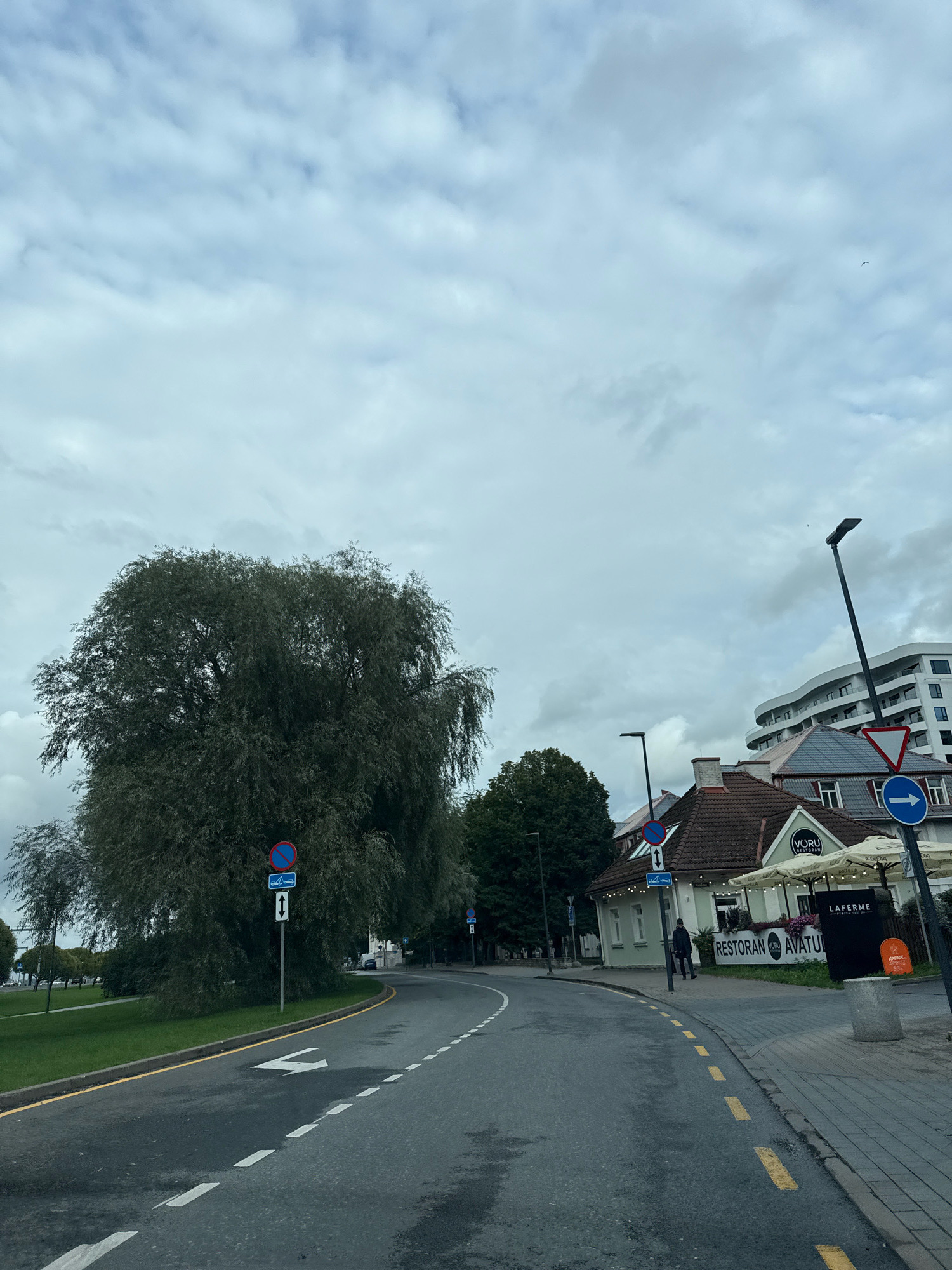 The image captures a street scene in Tallinn, Estonia, on a cloudy day. The road curves to the right, with lane markings indicating two lanes. A large, leafy tree dominates the left side of the frame, with a traffic sign showing a prohibition symbol and an arrow indicating two directions. On the right side of the road, a building with a tiled roof and an outdoor seating area houses "VÕRU RESTORAN" and "LAFERME." Several other buildings, including a modern, curved white structure, are visible in the background. A lamppost stands tall near the restaurant, and a blue directional arrow sign is mounted on a pole. A person is walking on the sidewalk in front of the restaurant. The environment appears to be a city street with sidewalks, roads, buildings, and trees.
