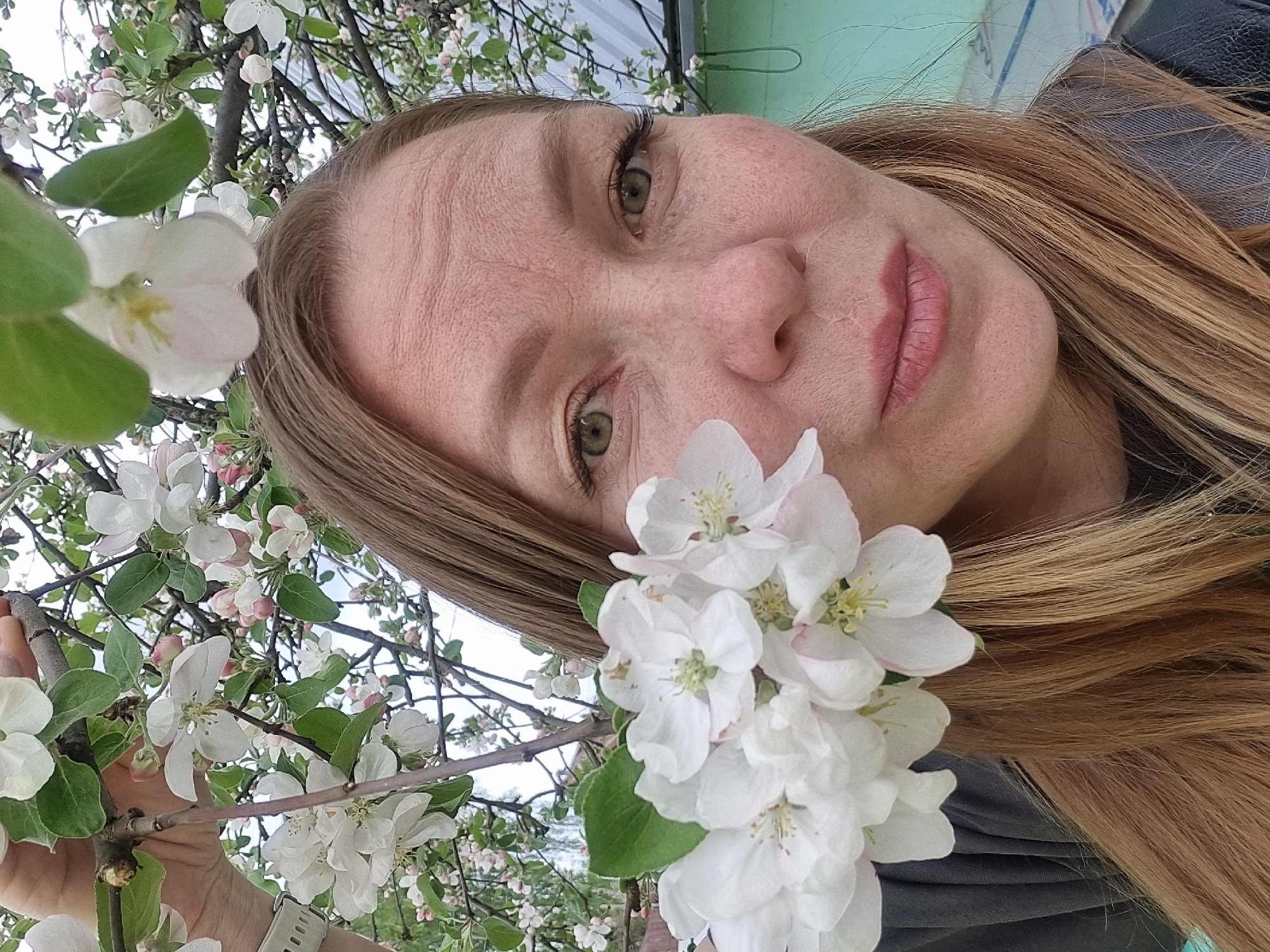 A woman with long, light brown hair and freckles poses for a selfie outdoors. She is surrounded by white flowers, possibly from an apple or cherry tree, with some of the blossoms held close to her face. The background includes hints of a light green wall or structure and a grey or silver corrugated metal surface. A hand can be seen holding a branch of the flowering tree, with a white watch visible on the wrist. The photograph's location is identified as Chaikovsky, Russia.