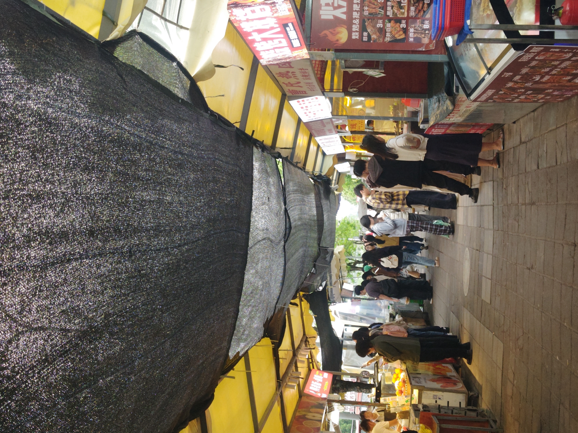 Here's a detailed description of the image:
**Environment:** The image depicts a street scene, likely a food market or a street with food vendors, in Shapingba District, China. The area is shaded by a combination of yellow awnings and black netting. The pavement is made of tiles.
**People:** There are many people walking and browsing the vendor stalls. They are dressed in casual clothing. They appear to be queuing or gathering around various stalls.
**Objects:** There are various food stalls with signs in Chinese, displaying prices and food options. There are also items on display, such as fruit.
**Actions and Interactions:** People are interacting with the food stalls. The general atmosphere suggests a busy commercial area.