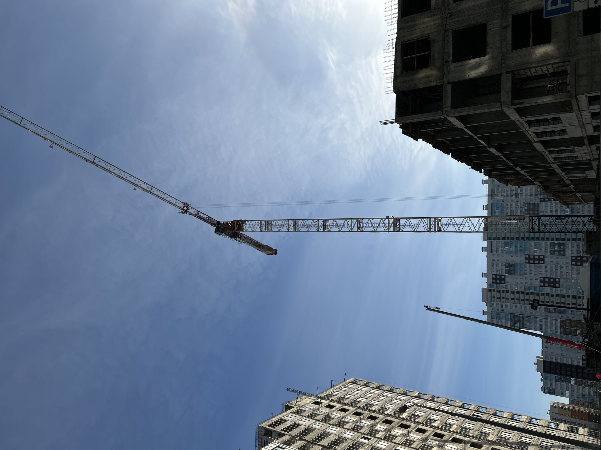 The image captures a construction site in Saint Petersburg, Russia, viewed from a low angle looking upwards. A tower crane, extending horizontally across the frame, dominates the scene. The crane's arm stretches from the upper-left to the center. Surrounding the crane are several multi-story buildings. Some are under construction, with unfinished facades and visible skeletal structures. Other buildings in the background appear to be completed residential or commercial structures, featuring a grid pattern of windows. The sky is partially cloudy, with a gradient from blue to white. No people are visible in the frame.