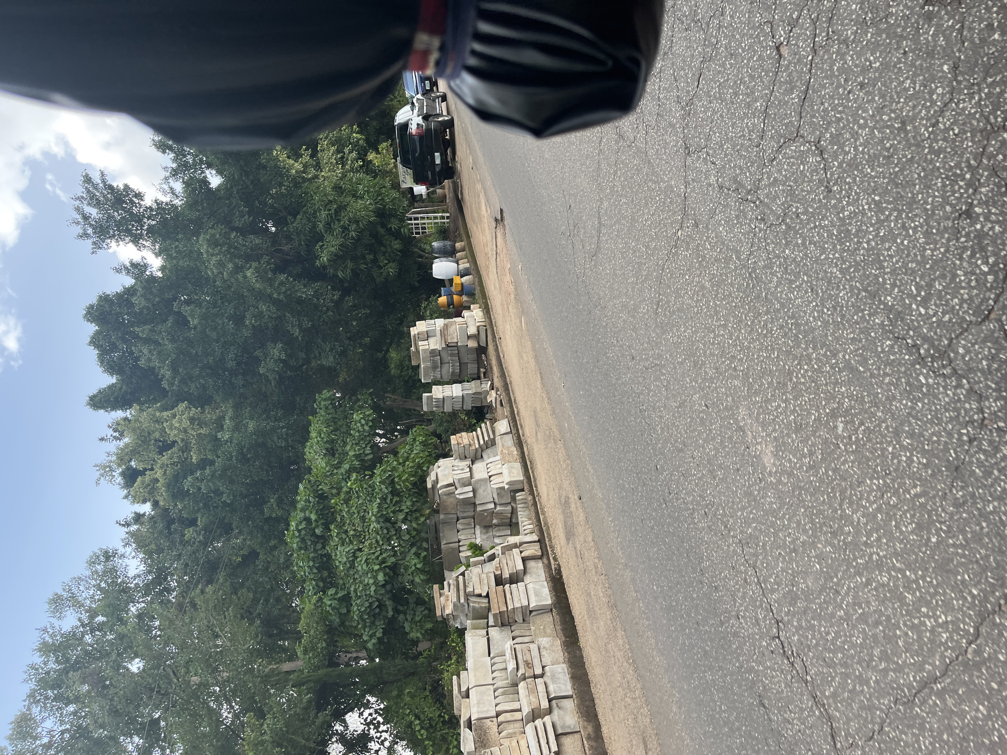 The image depicts a scene from Jos, Nigeria, likely during daylight hours given the brightness. The perspective seems to be from a vehicle, possibly looking sideways and slightly down.
In the foreground, a paved road with visible cracks is prominent. To the left of the road, stacks of concrete paving stones are arranged along the roadside, suggesting a construction or landscaping supply area. Further back, several vehicles, including a black SUV and what appears to be a truck, are parked or moving along the road. Beyond the vehicles, a dense line of lush green trees forms a backdrop. The sky above is a light blue with scattered white clouds. A person's shoulder and the edge of a dark, possibly leather, bag are visible at the very top of the frame, reinforcing the idea that the photo was taken from inside a moving vehicle. The overall impression is a snapshot of an everyday scene on a Nigerian road, with elements of commerce and urban greenery. There is no discernible text in the image.