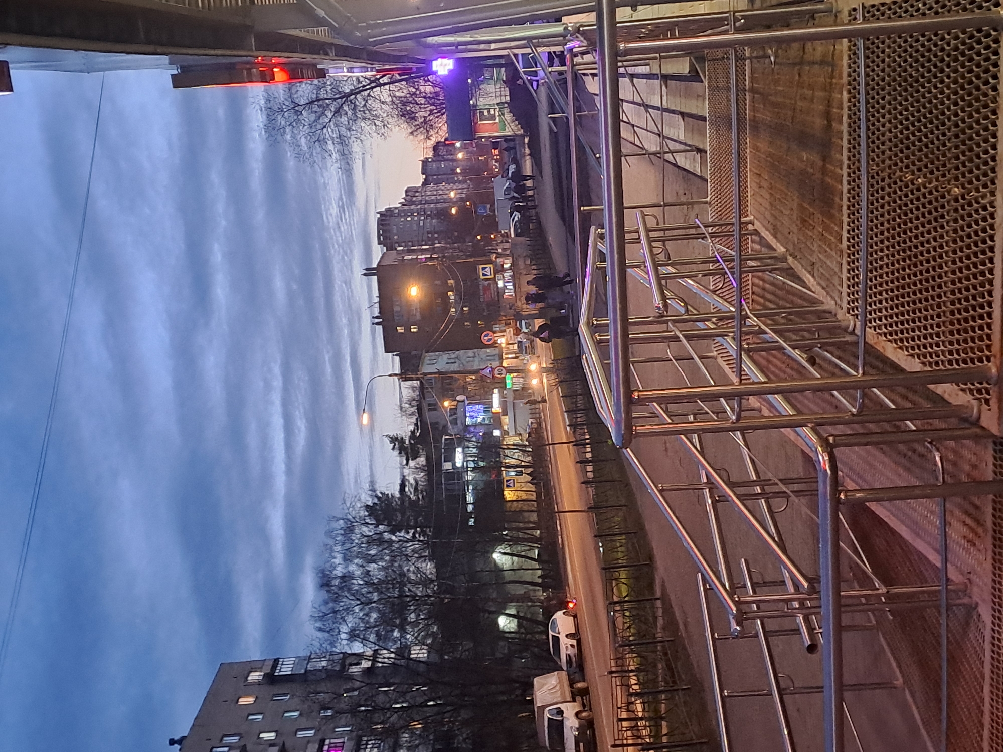 The image depicts a view of a street in Elektrostal, Russia, at dusk. The scene is framed by a metal railing structure in the foreground, possibly a staircase or barrier. In the background, multi-story apartment buildings line the street. Streetlights and building lights illuminate the scene, casting a warm glow on the roadway. Several cars are visible, along with a few pedestrians walking along the sidewalk. The sky is a mix of blue and gray, suggesting the transition from daylight to night. The environment appears urban, with a mix of residential and commercial buildings.