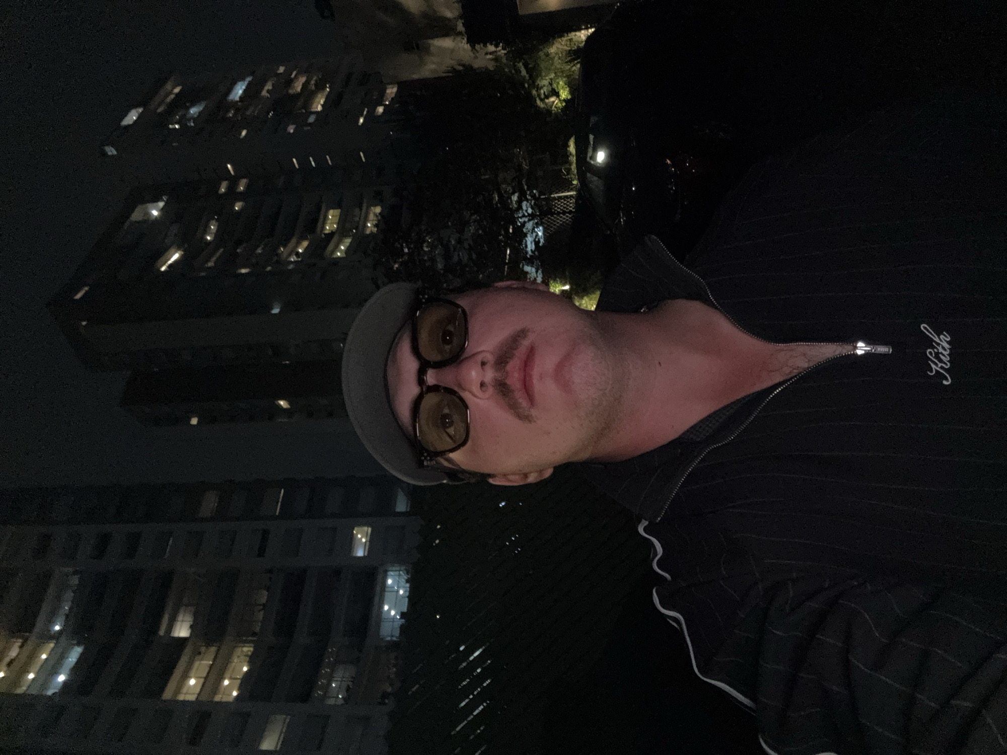 A person, likely male, is taking a selfie at night. The background includes buildings with illuminated windows, suggesting a city location. The person is wearing a hat and glasses and a dark zip-up jacket with the word "Kith" embroidered on it. The image appears to be taken from a high vantage point, possibly a balcony or rooftop. It is not possible to confirm the location is Huixquilucan de Degollado, Mexico, based on the visual information provided.