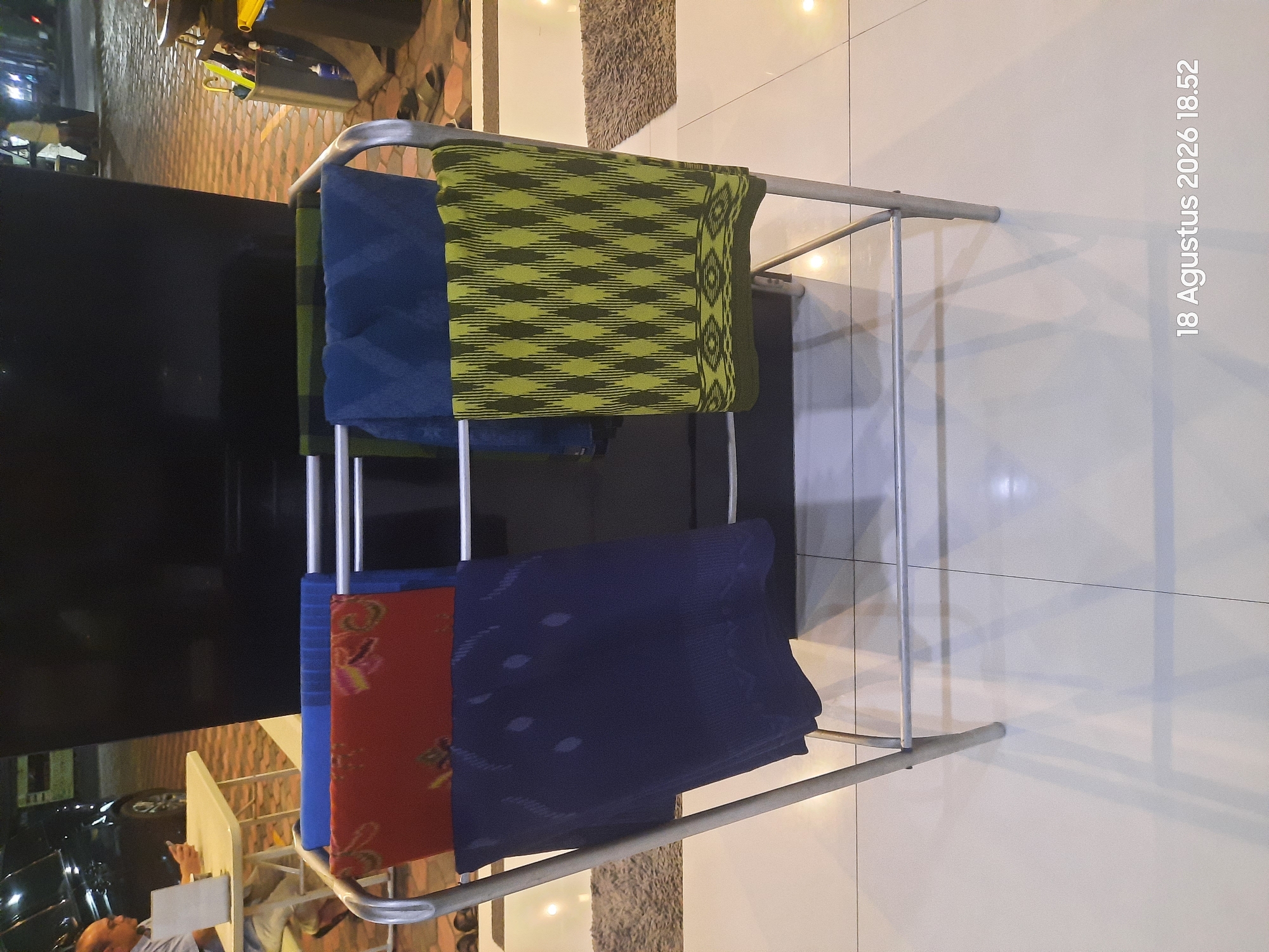 In Sukokerto, Indonesia, a man sits at a table on a patio, looking at his phone. In front of him is a drying rack with several pieces of fabric. The fabric pieces are stacked vertically on the rack. The top piece is red with a floral pattern. Below that is a dark blue piece with a dotted pattern. The next piece is a greenish-yellow fabric with a diamond pattern. The rack itself is made of metal and has a modern design. The patio floor is tiled with white tiles. In the background, a black rectangular object is visible, possibly a large screen or panel. There is also a vehicle parked nearby, and a trash bin. The lighting suggests it is evening or night.