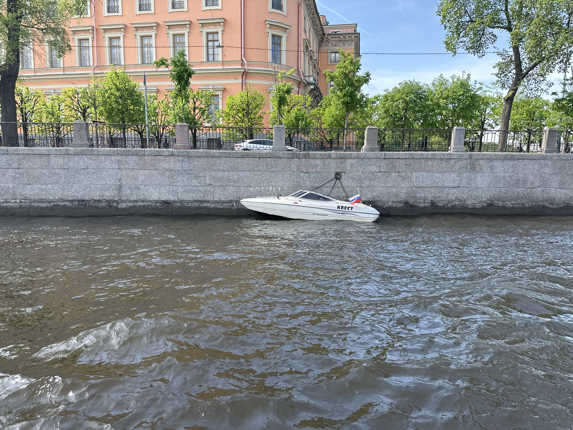 The image depicts a white motorboat moored to a granite embankment in Saint Petersburg, Russia. The boat bears the word "КВЕСТ" (Quest) in Cyrillic script and a Russian flag. The water appears murky with ripples. Behind the embankment is a metal fence, trees with green foliage, and a large peach-colored building. A car can be seen parked on the street behind the fence. The sky is partly cloudy.