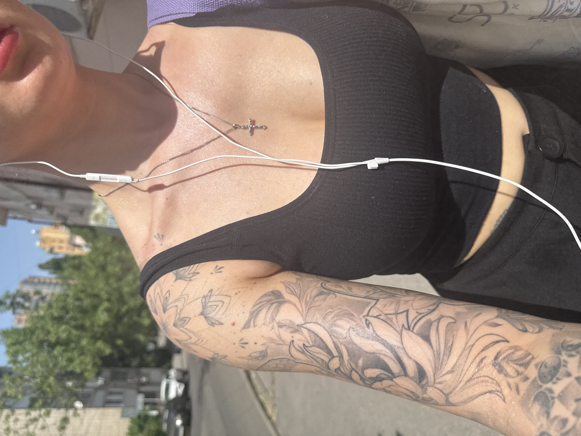 The image is a close-up, slightly angled view of a person outdoors. The person has fair skin and is wearing a black ribbed tank top. Part of a purple top is visible at the top of the frame. A silver cross necklace is worn around the neck. White wired headphones are also visible. The person has tattoos on their arm. The background shows green trees, buildings, parked cars and a street. Based on metadata, the location is Rostov-on-Don, Russia.