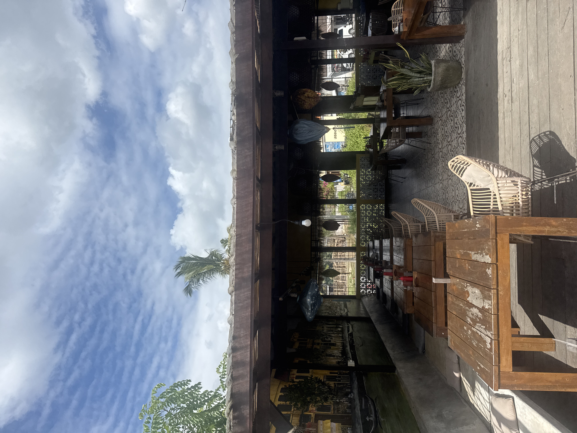 The image depicts an outdoor restaurant or cafe area with wooden tables and chairs. A thatched-roof structure with wooden beams provides shade. A mural adorns the wall on the left side of the structure, featuring a scene with buildings and a boat. Several decorative hanging lights are suspended from the ceiling. In the background, foliage and a glimpse of a road with a white car are visible. The restaurant appears to be located in Nusa Penida, Indonesia, given the tropical vegetation and architectural style. The sky is cloudy with patches of blue. The floor is a combination of wooden planks and patterned tiles. There are no people visible in the image.
