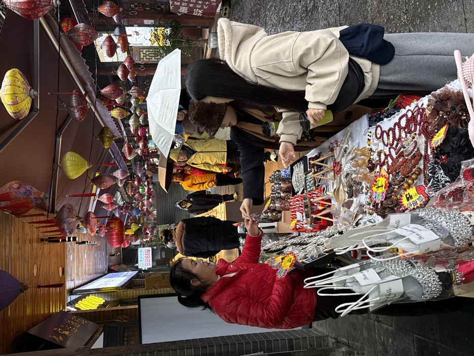 The image depicts an active outdoor market scene on a wet, paved street or alleyway within Pengshui Miao and Tujia Autonomous County, China. The overhead area is extensively adorned with numerous colorful traditional Chinese lanterns of varying shapes and sizes, suspended from wires and architectural elements.
In the foreground, a female vendor, appearing middle-aged and wearing a bright red puffer jacket, stands behind a white-topped stall displaying a diverse range of merchandise. She is engaged in an interaction with two younger women, presenting or handing a small, silver-colored item to one of them. The stall items include hanging silver-colored ornaments, small containers with colorful contents, red-and-white striped objects, wooden spoons, carved figurines, multiple beaded bracelets (predominantly red and brown hues), and various hair accessories. Several white paper bags containing silver-colored items are visible to the vendor's left.
The two female customers stand to the right of the stall. One woman, with long dark hair, wears a light beige hooded jacket, grey sweatpants, and a black fanny pack, holding a clear cup with a green beverage. The other woman, partially obscured, wears a dark coat, a brown furry hat, and also holds a clear cup with a green beverage.
In the background, the street inclines uphill, transitioning into stone steps. Multiple pedestrians are visible, some walking with open umbrellas (one prominent white umbrella, and several darker ones), indicating wet conditions, likely from recent or ongoing rain. Individuals in the background are dressed in various casual outerwear, including a bright yellow jacket and dark puffer jackets. Buildings with traditional Chinese architectural features, including wooden elements, line both sides of the thoroughfare. An illuminated sign with yellow characters is visible on a building to the left, and a traditional wooden doorway and foliage are seen to the right.