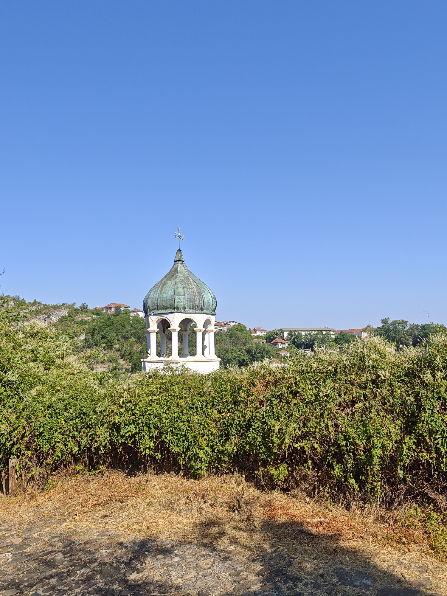 The image shows a bell tower with a green dome and a cross on top, situated on a hill overlooking a city. The bell tower is surrounded by lush green trees and bushes. In the background, there are several buildings with red-tiled roofs, typical of Lovech, Bulgaria. The sky is clear and blue. The foreground consists of a cobblestone path and dry grass. The scene appears to be a sunny day in a historical city.