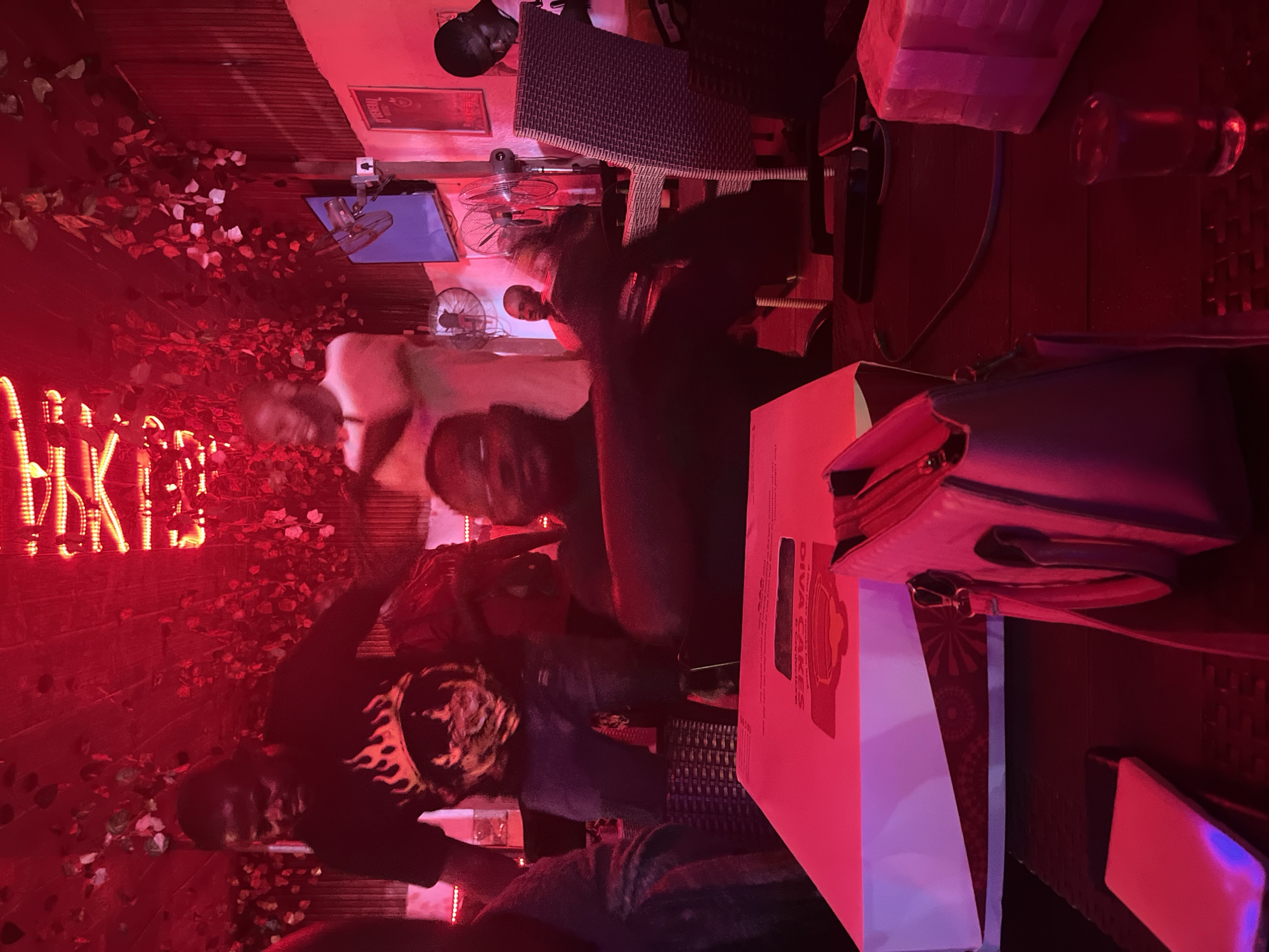 A slightly blurred, low-lit photograph taken in an indoor establishment in Ado, Nigeria, depicts at least three people at a table. The light is predominately red. One person wears a black shirt with a fire design on one sleeve. At least two people are seated and are looking at the camera. The table has a Diva Cakes box on it. A purse sits to the right of the cake box on the table. A neon sign is on the left. There are small electric fans attached to the wall. Other tables and chairs are in the background. A glass of liquid is on the right.