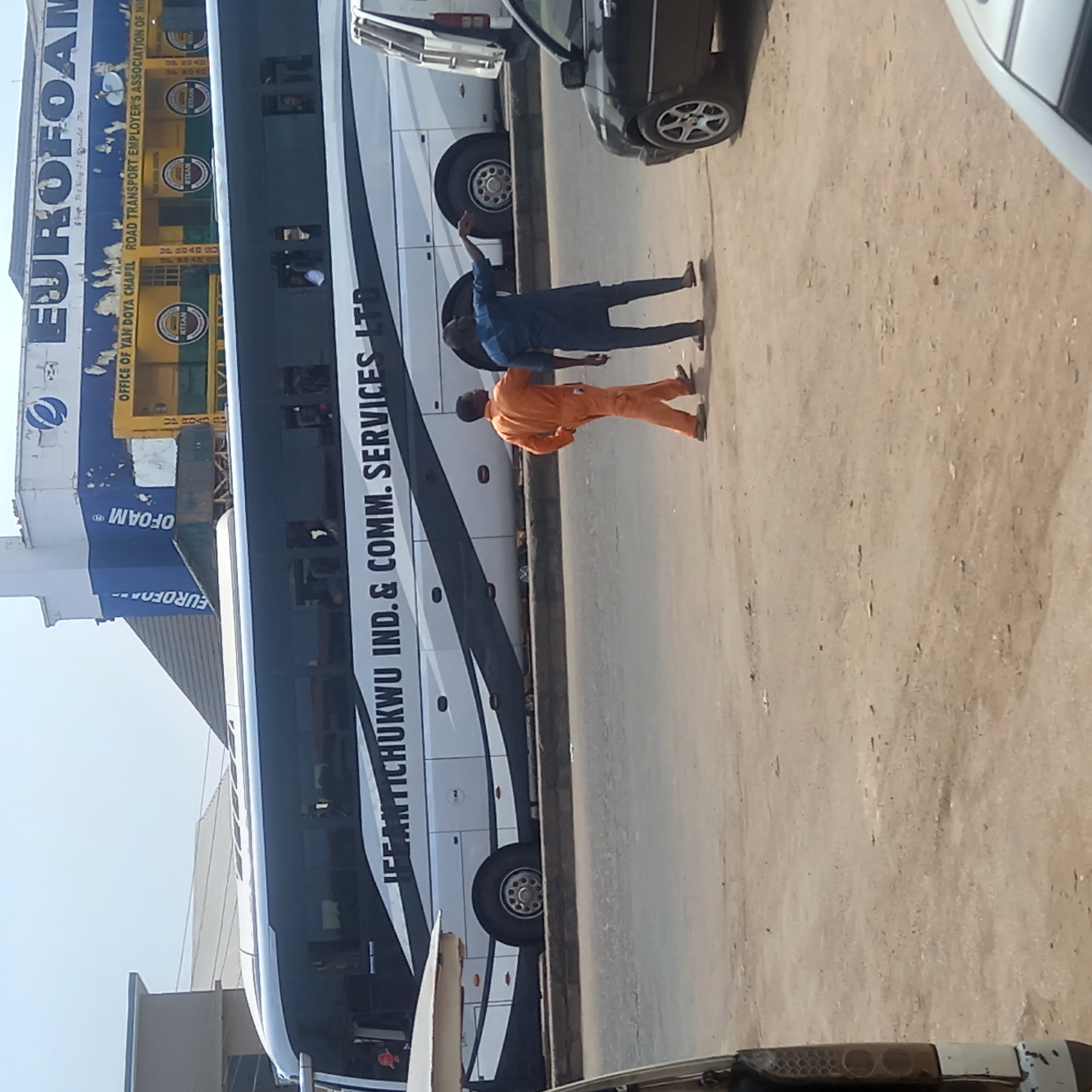 The image depicts a bustling street scene in Dadi Riba, Nigeria, under bright daylight. A long, white and black passenger bus, prominently displaying "IFFANTICHUKWU IND. & COMM. SERVICES LTD" along its side, is parked parallel to a building on the left. Several windows of the bus show hints of people inside.
Behind the bus, a commercial building with a faded blue and yellow exterior features multiple signs. Large lettering spells out "EUROFOAM," and detailed text identifies it as the "OFFICE OF YAN DOYA CHAPEL ROAD TRANSPORT EMPLOYER'S ASSOCIATION OF NIC." Several "RTEAN" logos are also visible, along with phrases like "Stop Shiny 3V Would Do" and "UP ROADS."
On the sandy-colored ground in front of the bus, two men are captured in interaction. One man wears a bright orange jumpsuit, facing away from the viewer towards the bus. Beside him, another man, dressed in a blue traditional garment, has his arm raised in a gesture, possibly addressing the other man or pointing towards the bus.
Parts of other vehicles, including a black car on the right and a white car further right, are visible. The ground appears to be a dusty or sandy surface, common in urban areas. The bright lighting suggests a clear, sunny day.