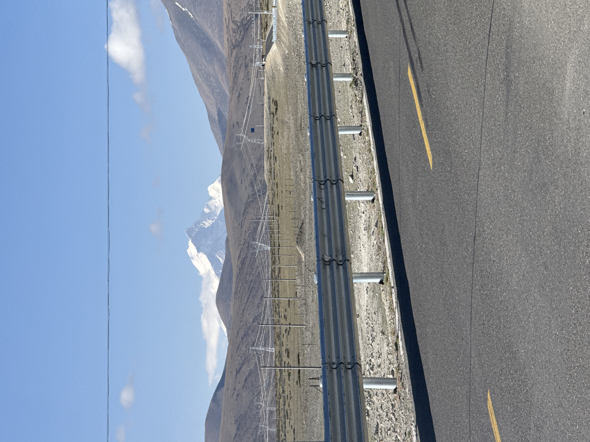 The image presents a vast, high-altitude landscape in Gyirong County, China, under a clear, sunny sky. The scene is dominated by a combination of natural rugged terrain and human infrastructure.
In the foreground, a paved asphalt road occupies the right side of the frame, featuring a yellow dashed line marking its center. To the left of the road, a robust metal guardrail separates the roadway from a rocky, gravelly embankment.
Beyond the guardrail and embankment, the midground opens into a wide, dry plain or valley. This terrain appears arid and sparsely vegetated, with light-colored soil and rocks. Stretching across this expanse are numerous electricity transmission towers and power lines, receding into the distance and indicating significant infrastructure.
The background is filled with majestic mountains. Those closer to the viewer exhibit a rugged, rocky appearance, while a prominent peak further away stands out with a distinct snow cap and visible snowfields, suggesting high elevation. The sky above is a vivid blue, with a few scattered white clouds. The strong, direct lighting suggests it is daytime, likely morning or afternoon.
No people or specific activities are depicted in the image, presenting a static, grand landscape. There is no visible text. The overall impression is one of a remote, mountainous region with a blend of natural grandeur and utilitarian development.