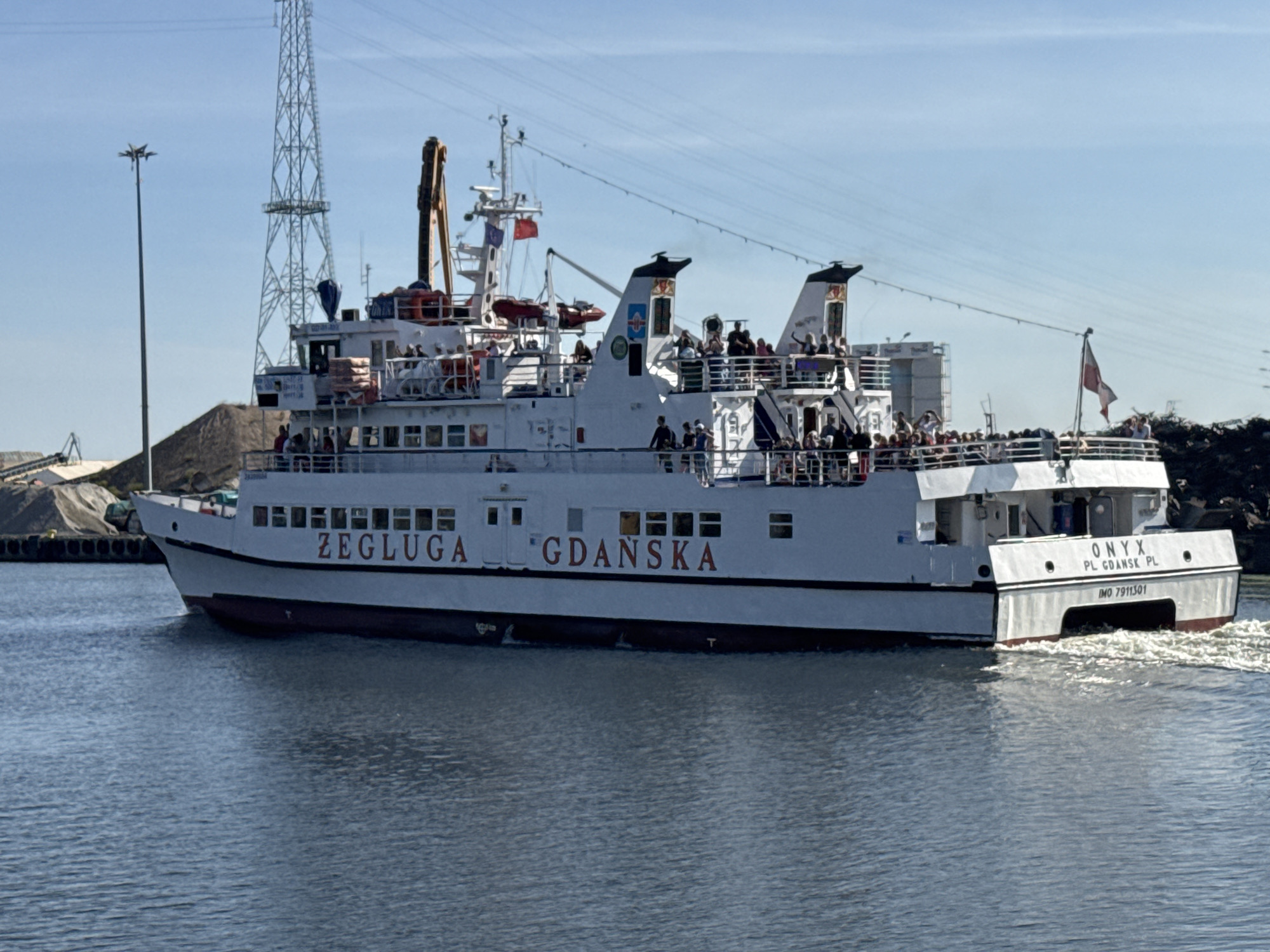 This is a medium shot, eye-level, exterior, daytime, medium-angle photograph of a white ferry boat named "ONYX" with red lettering. The ferry is in motion on a body of water, creating a wake behind it. The boat has multiple decks with people visible on them, many looking out at the water. The side of the ferry is labeled "ŻEGLUGA" and "GDAŃSKA" in red lettering. The rear of the boat shows the name "ONYX" and "PL GDANSK PL" above the registration number "IMO 7911301". In the background, a large industrial tower with a power line crosses behind the ferry. To the left of the ferry, a large pile of dark gravel or coal is visible, with industrial equipment and a conveyor belt. To the right, there is a rocky, dark shoreline or industrial debris. The sky is clear and blue. The overall impression is an industrial waterfront scene in Gdańsk, Poland.