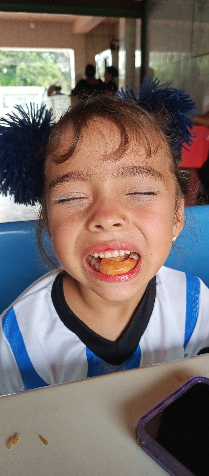 A young girl with brown hair and blue hair accessories is pictured indoors with her eyes closed and a piece of food in her mouth. She is wearing a white shirt with blue stripes and a black collar. A cell phone and some scattered pieces of food are visible on a table in the foreground. The background suggests a dining area with other people present. The location is identified as Tinaquillo, Venezuela.